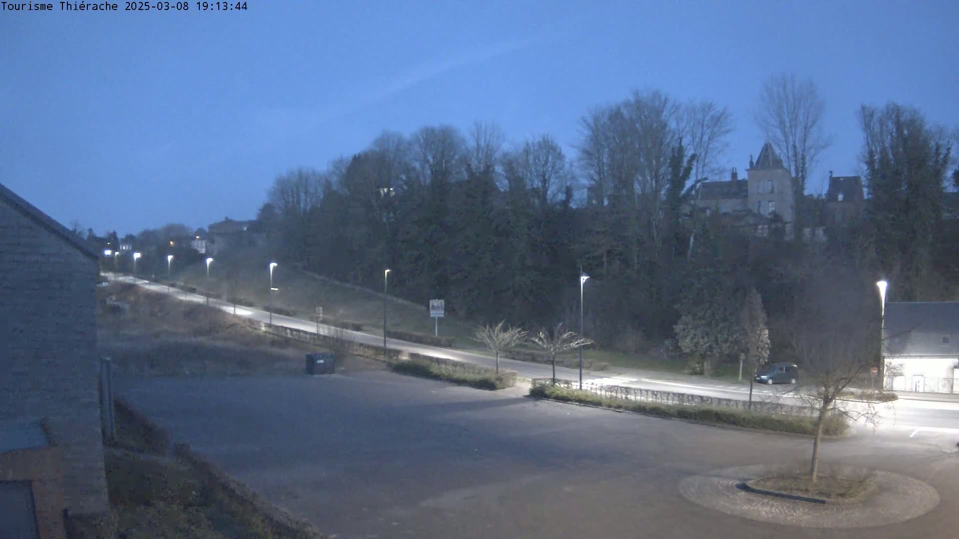 An empty parking lot at dusk, with a road leading uphill towards a hill covered in trees and buildings.