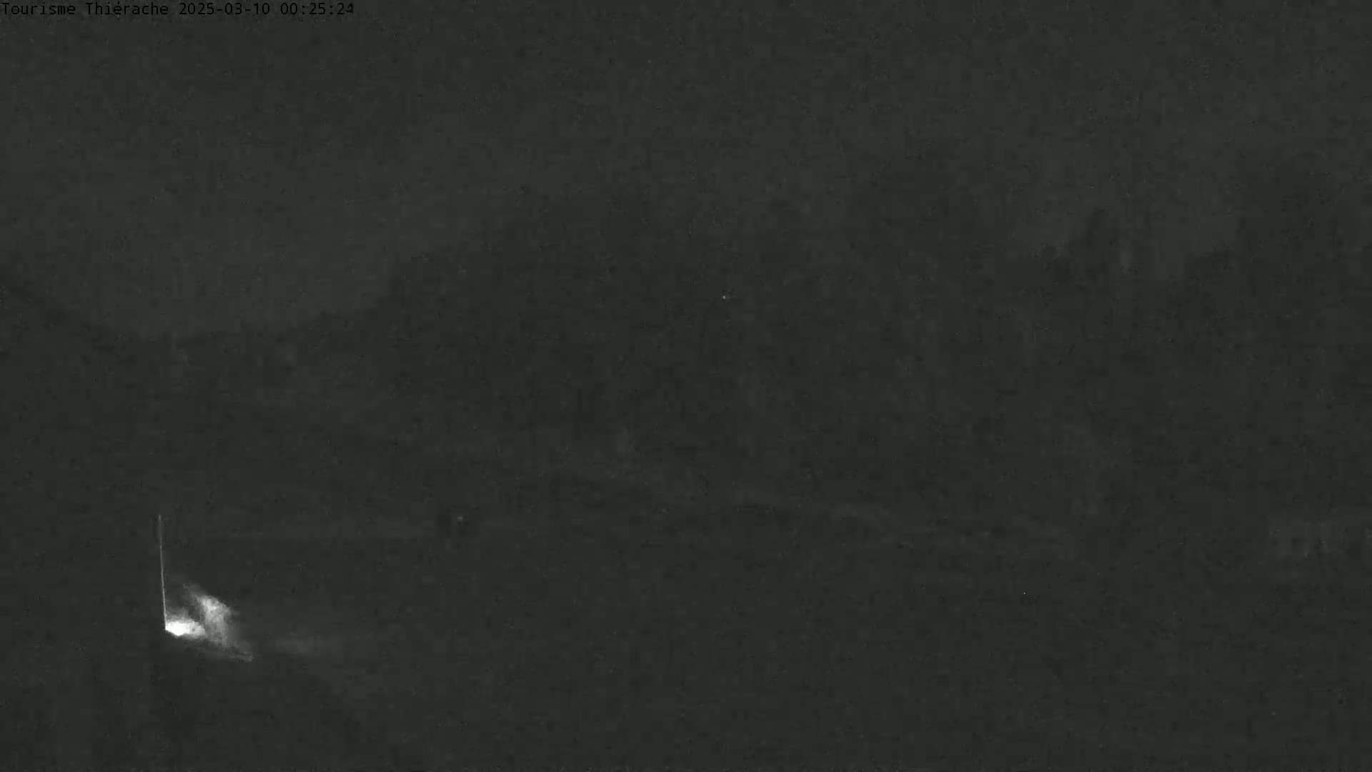 A dark, nighttime scene shows a dimly lit area with indistinct shapes in the background under a cloudy sky.