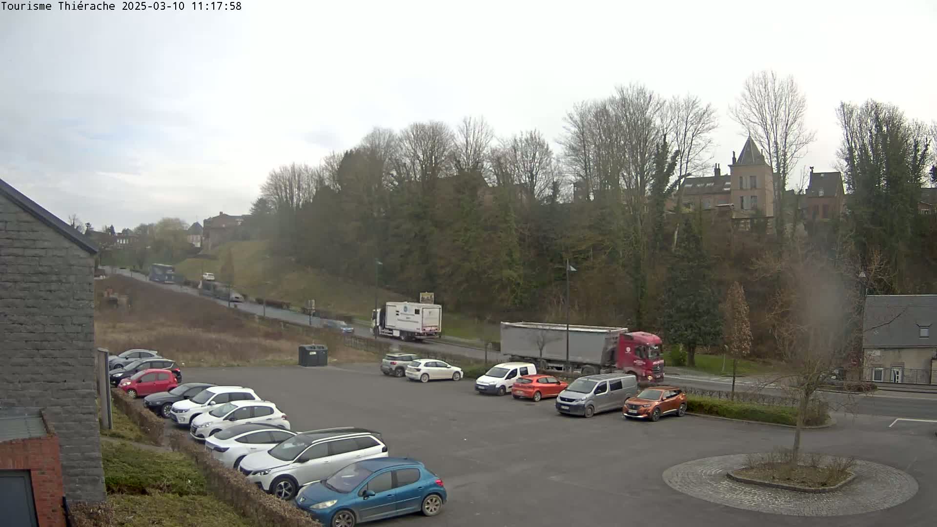 A parking lot with several cars is situated below a hill covered in bare trees, beyond which sits a village with buildings and a prominent tower under an overcast sky.