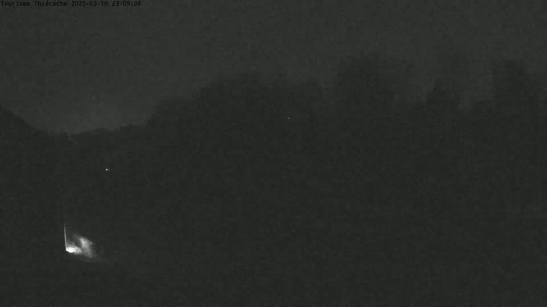 A dark, nighttime outdoor scene shows a dimly lit area with some trees in the background under a cloudy sky.