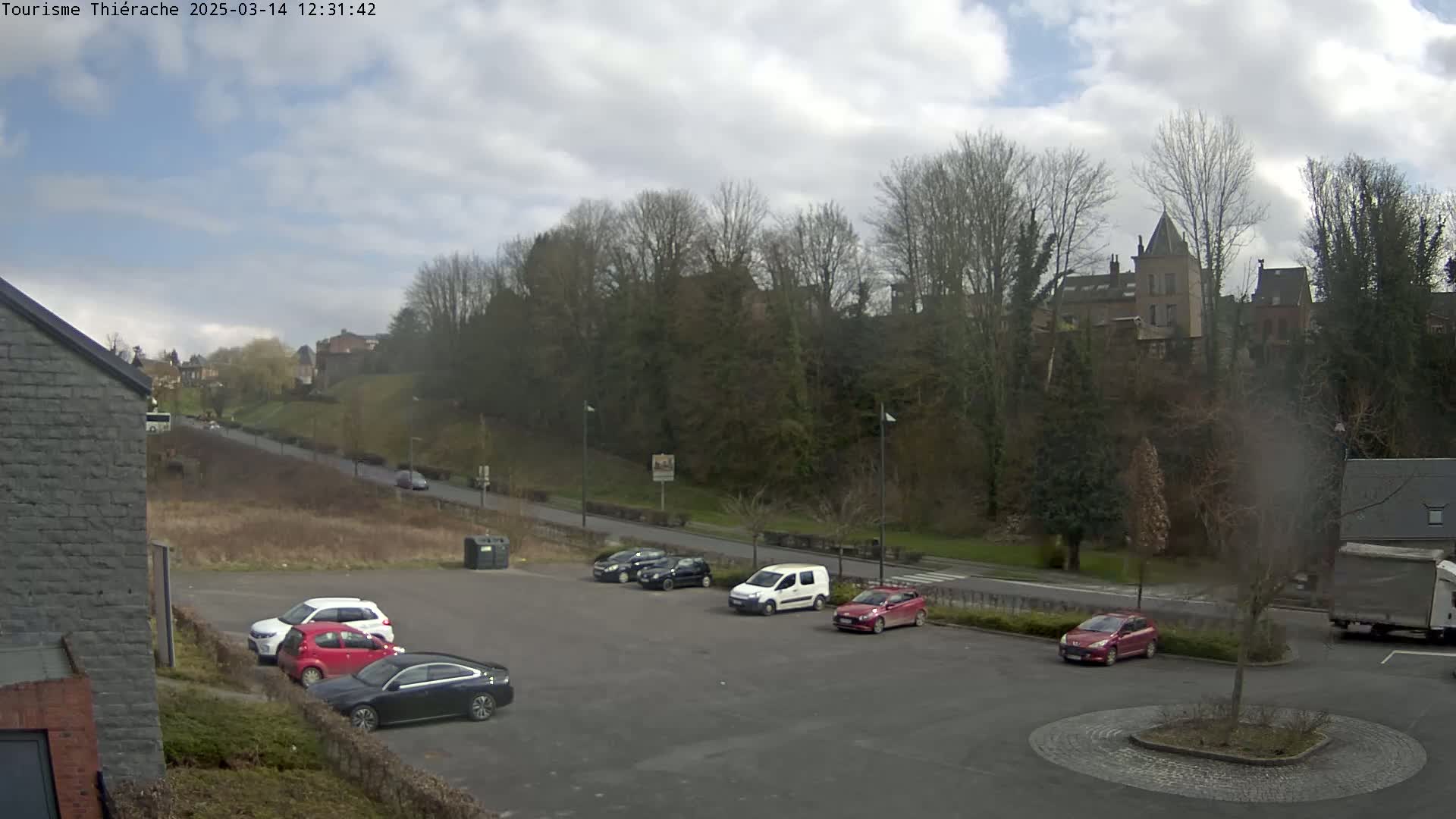 A parking lot with several cars is situated below a hill with trees and buildings under a partly cloudy sky.