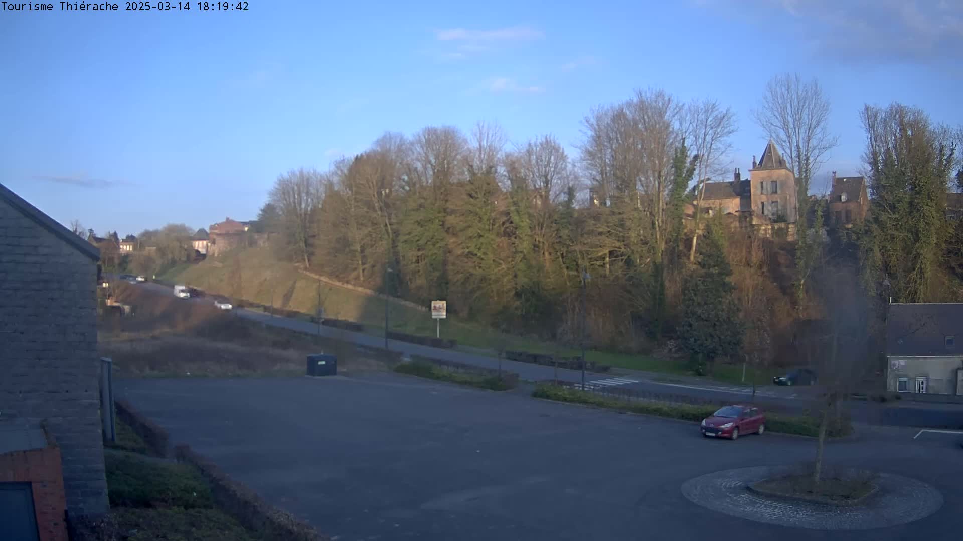 A mostly empty parking lot overlooks a road leading to a hill covered in leafless trees and topped with a stone building under a clear blue sky.