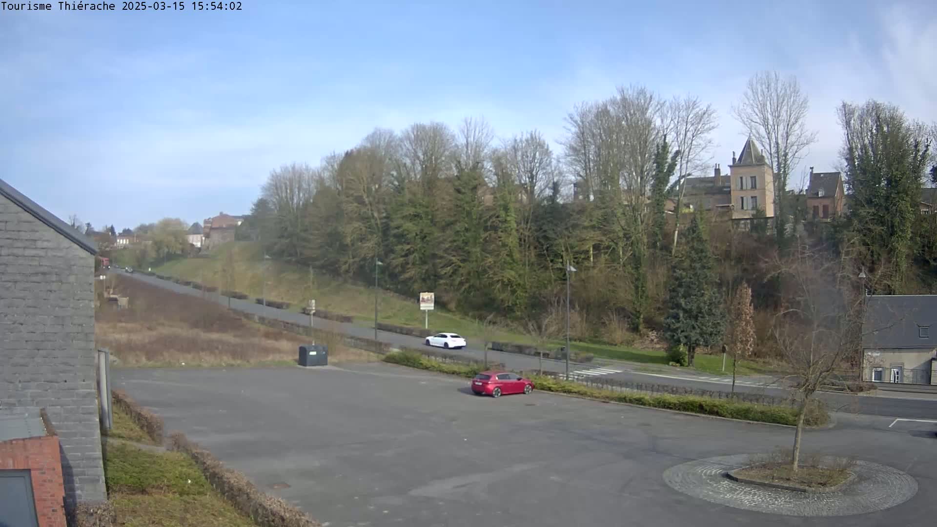 A mostly empty parking lot with two cars, one red and one white, overlooks a road and a hillside with bare trees and buildings in the distance under a clear, sunny sky.