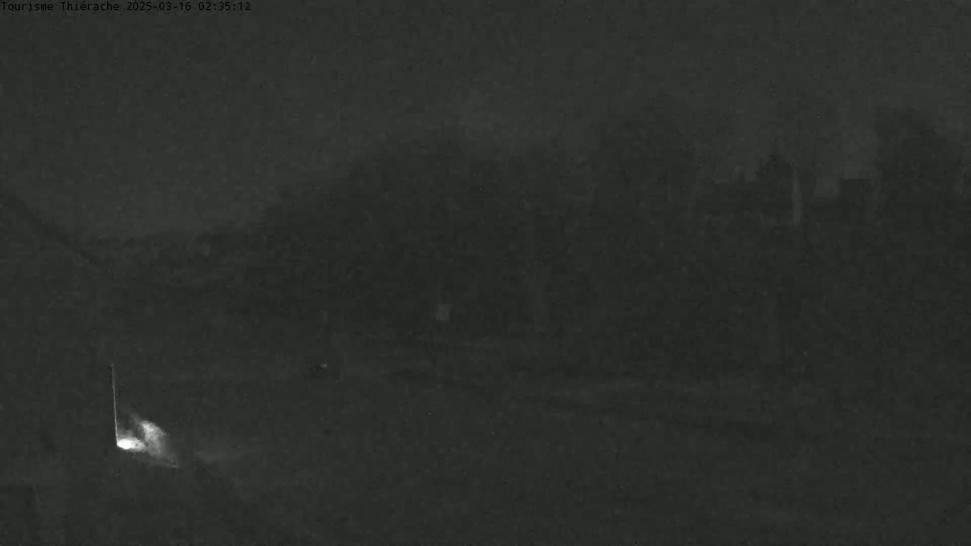 A low-light, nighttime image shows a dark, blurry landscape with indistinct shapes in the background and a small, bright light source near the bottom left corner.