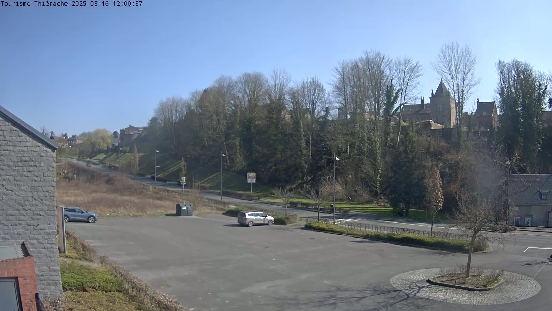 A mostly empty parking lot overlooks a road leading to a hillside covered in leafless trees and buildings under a clear, sunny sky.