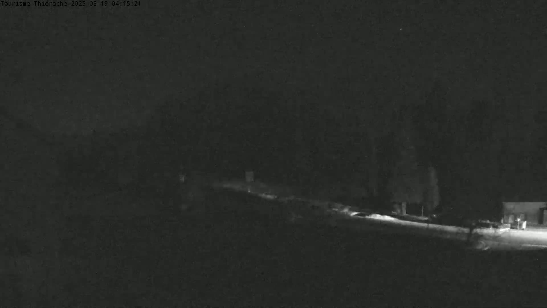 A dark, nighttime scene shows a snow-covered road curving through a sparsely wooded area under a clear sky.