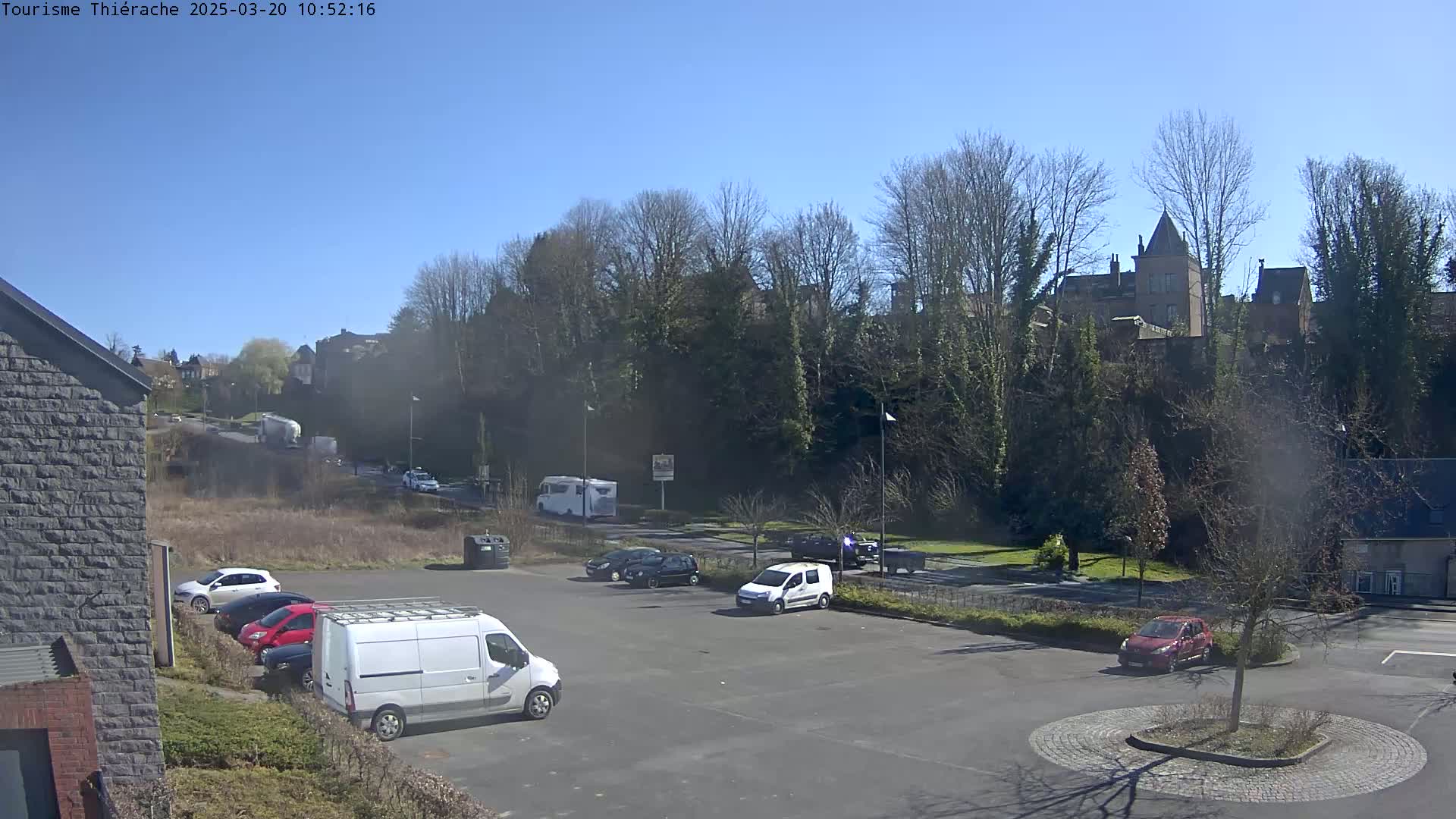 A sunny day shows a parking lot with several cars and a van, situated near a road and a line of trees overlooking buildings on a hill.