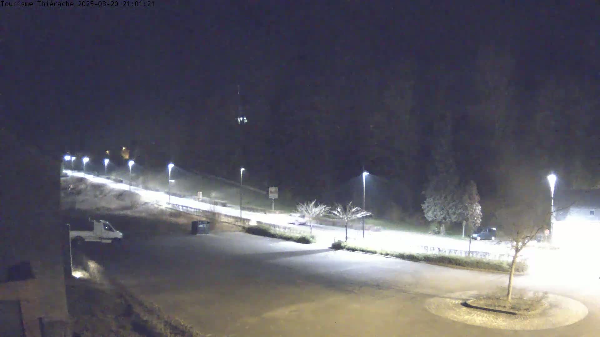 A nighttime view of a mostly empty parking lot illuminated by streetlights, with a few vehicles present and a road leading up a hill into a dark wooded area.