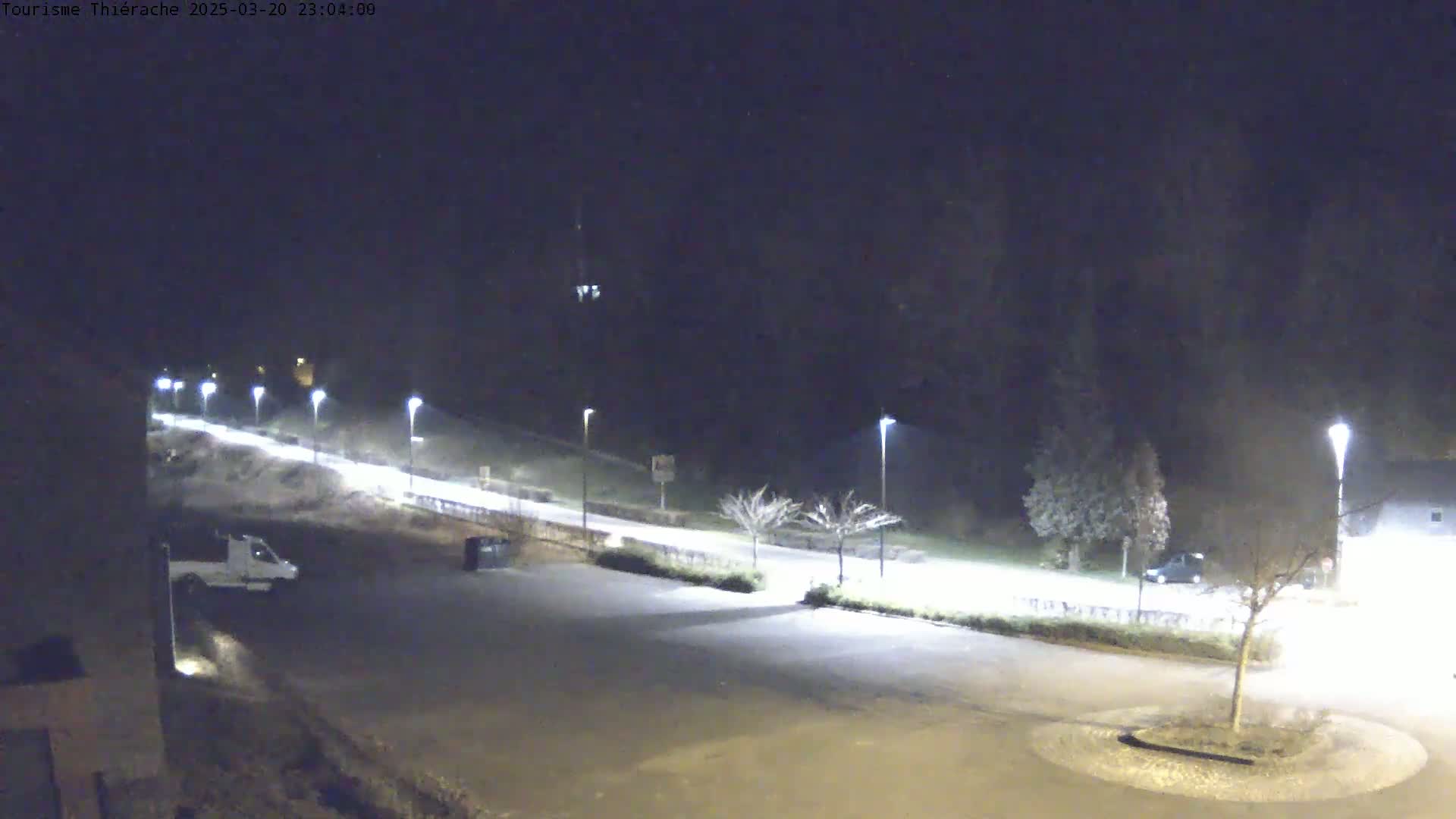 A nighttime, dimly lit parking lot and road are shown, with a few vehicles present and trees sparsely lining the road.