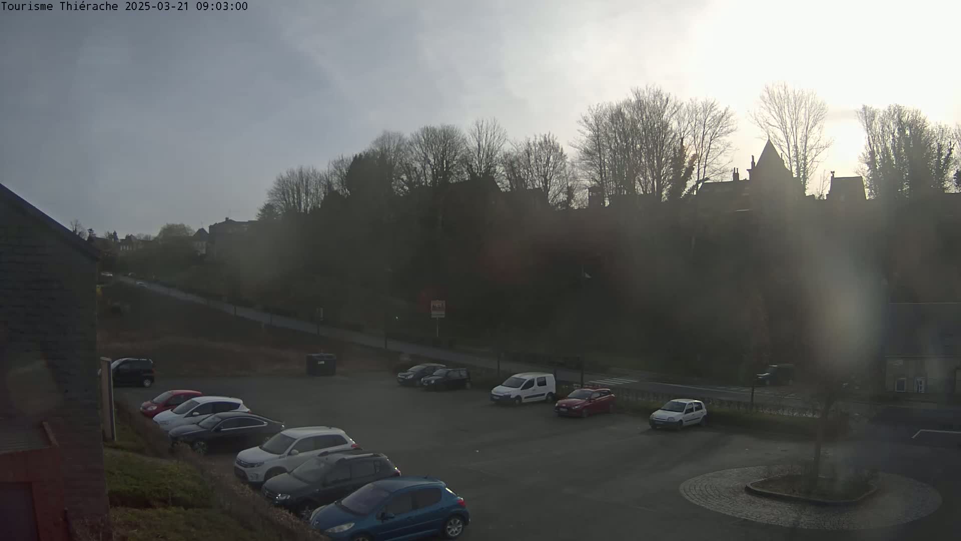 A hazy, overcast day reveals a parking lot with several cars, situated below a hill topped with buildings and trees.