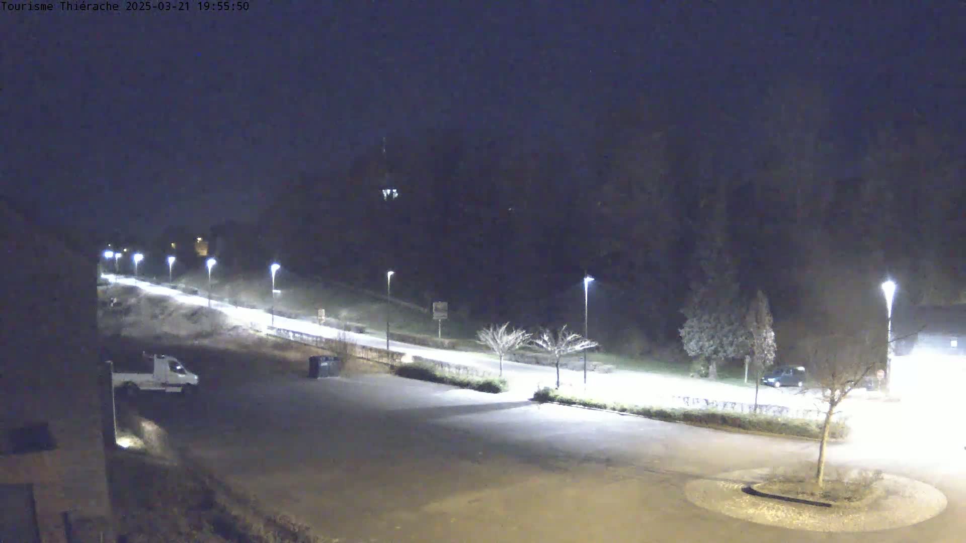 A nighttime, dimly lit parking lot and road are visible, with a few vehicles and trees sparsely scattered throughout.