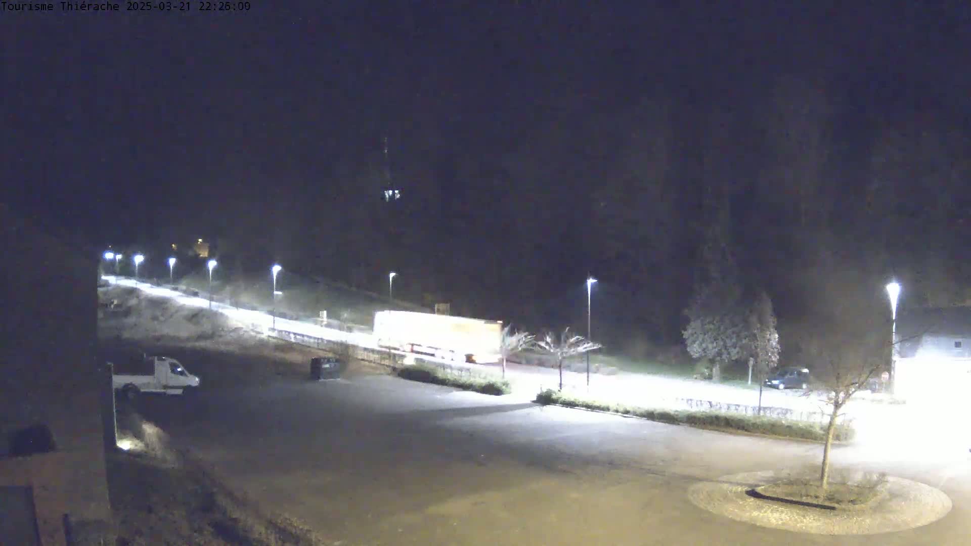 A nighttime view of a mostly empty parking lot with a few vehicles, illuminated by streetlights, next to a road with some other vehicles driving on it.