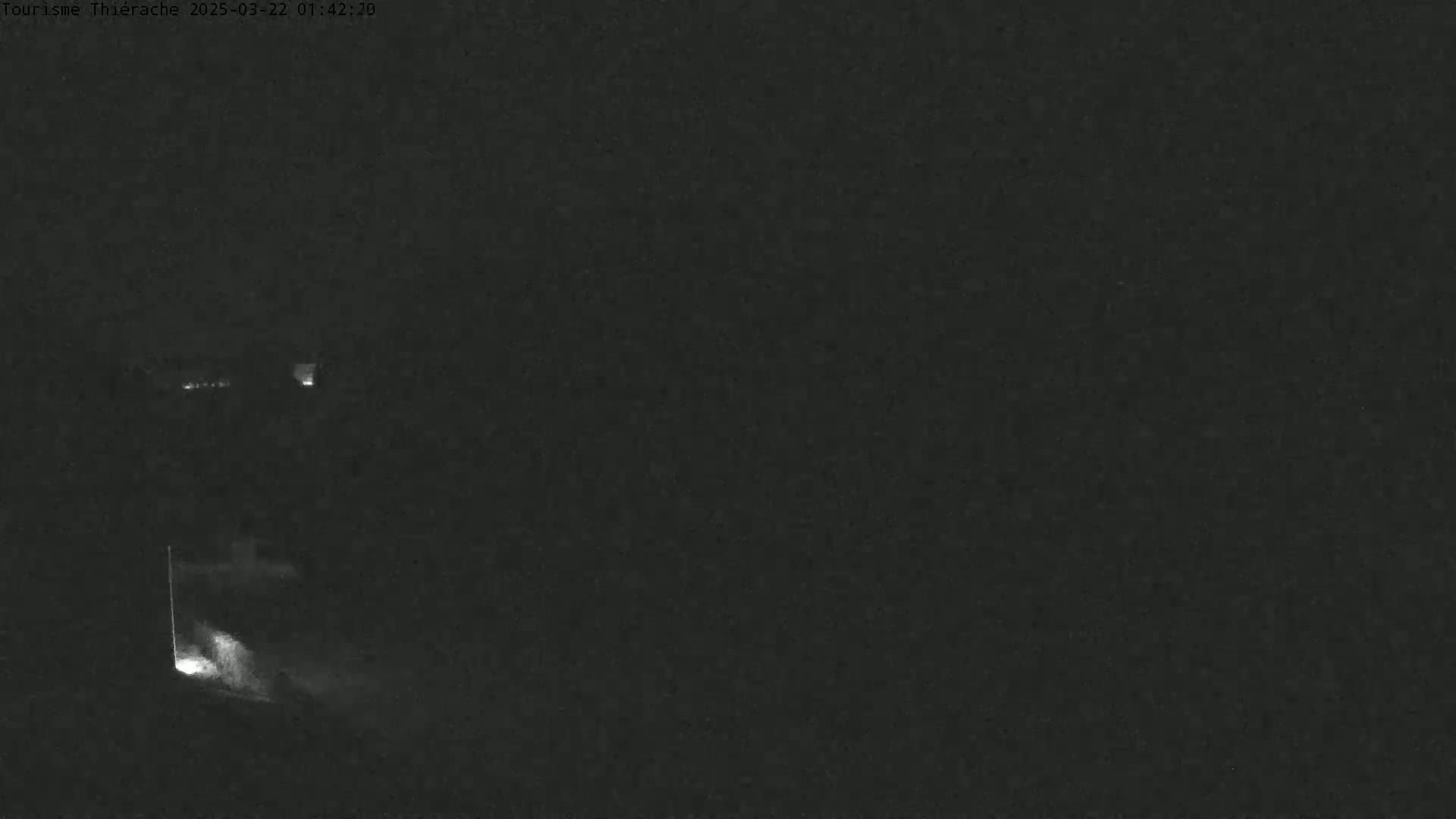 A nighttime, low-light image shows a dark, possibly wooded area with a faint light source emanating from a structure or opening in the lower-left.