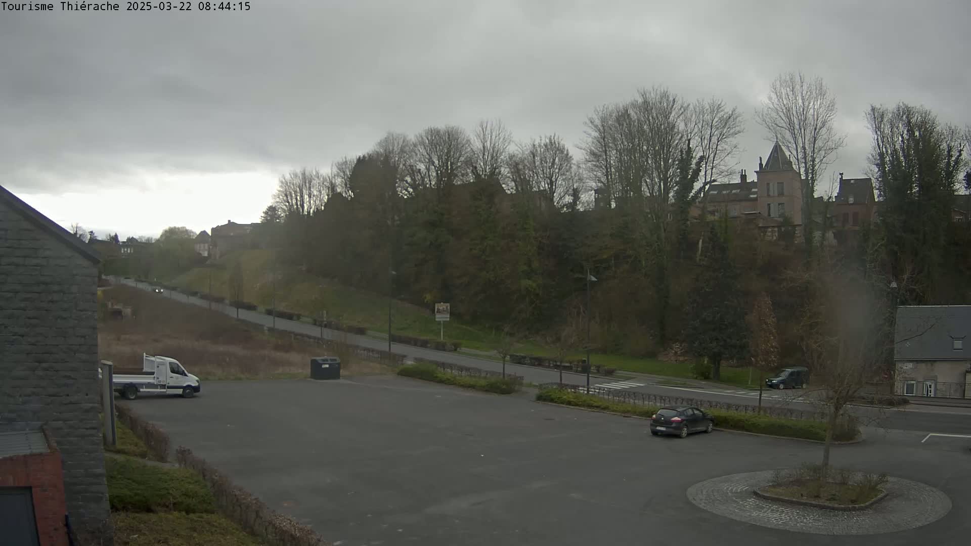 A mostly empty parking lot with a few vehicles, a white van and a dark-colored car, overlooks a road leading to a hill covered in leafless trees and buildings under an overcast sky.