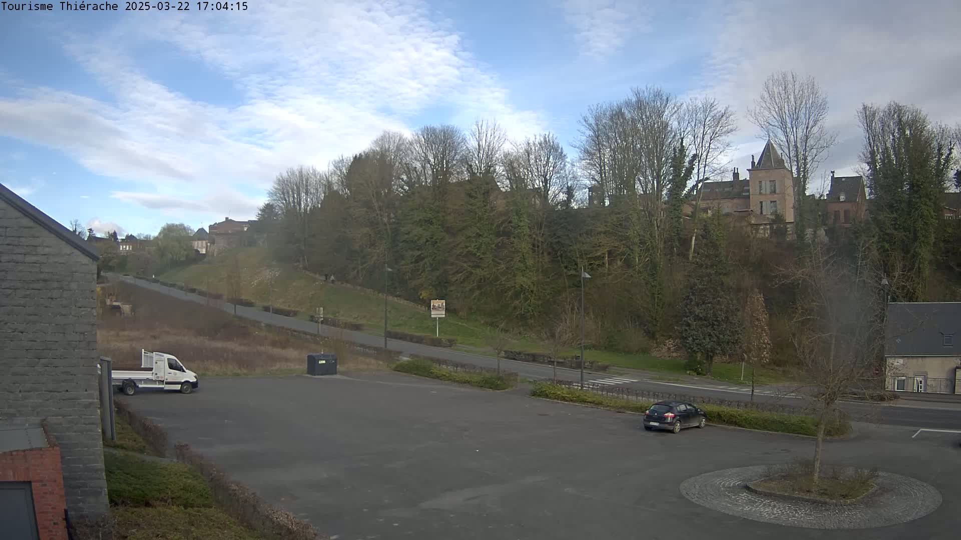 A mostly clear day shows a parking lot with a few vehicles, next to a road that leads up to a hillside with buildings and trees.