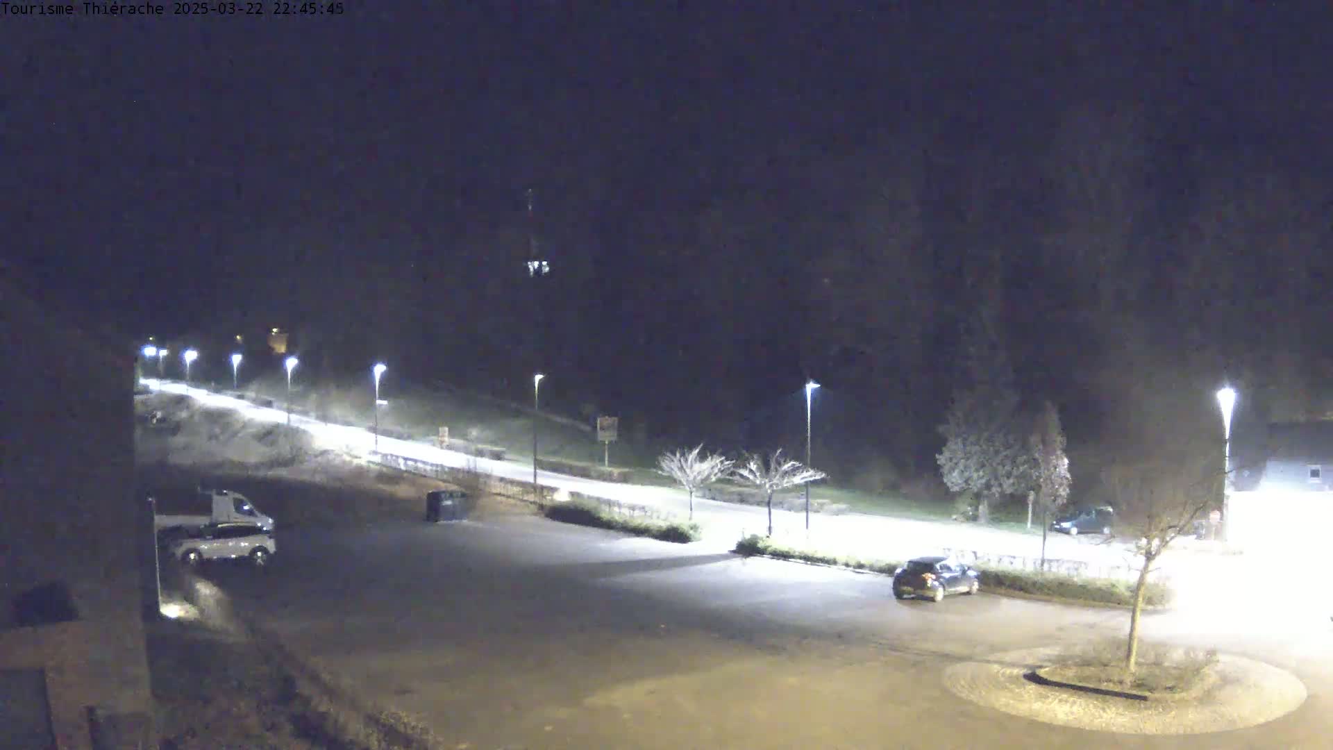 A nighttime view of a mostly empty parking lot next to a road lit by streetlights, with a few cars parked and some sparse trees visible.