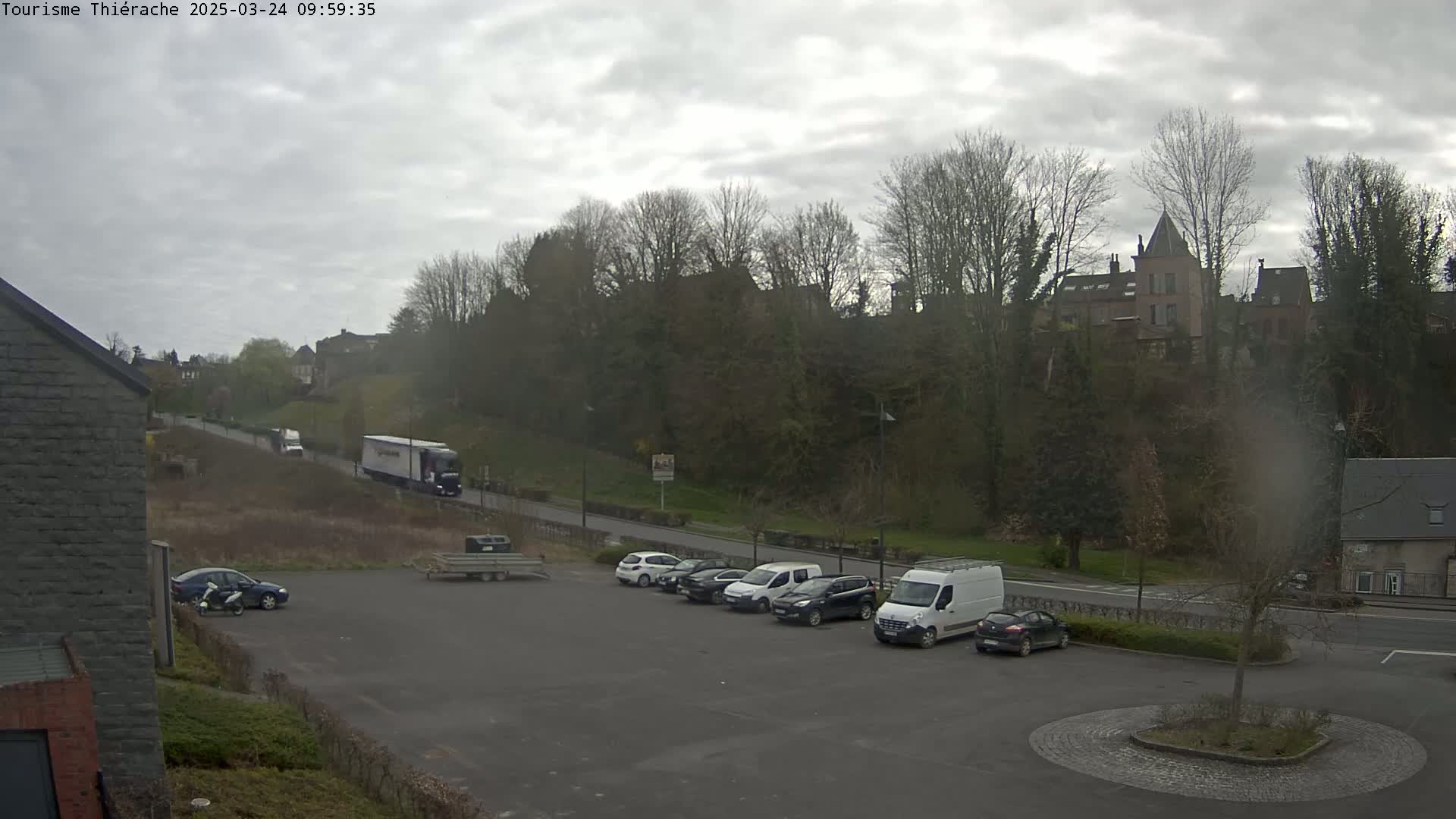A parking lot with several cars and a van, situated in front of a hill with buildings and trees under an overcast sky.