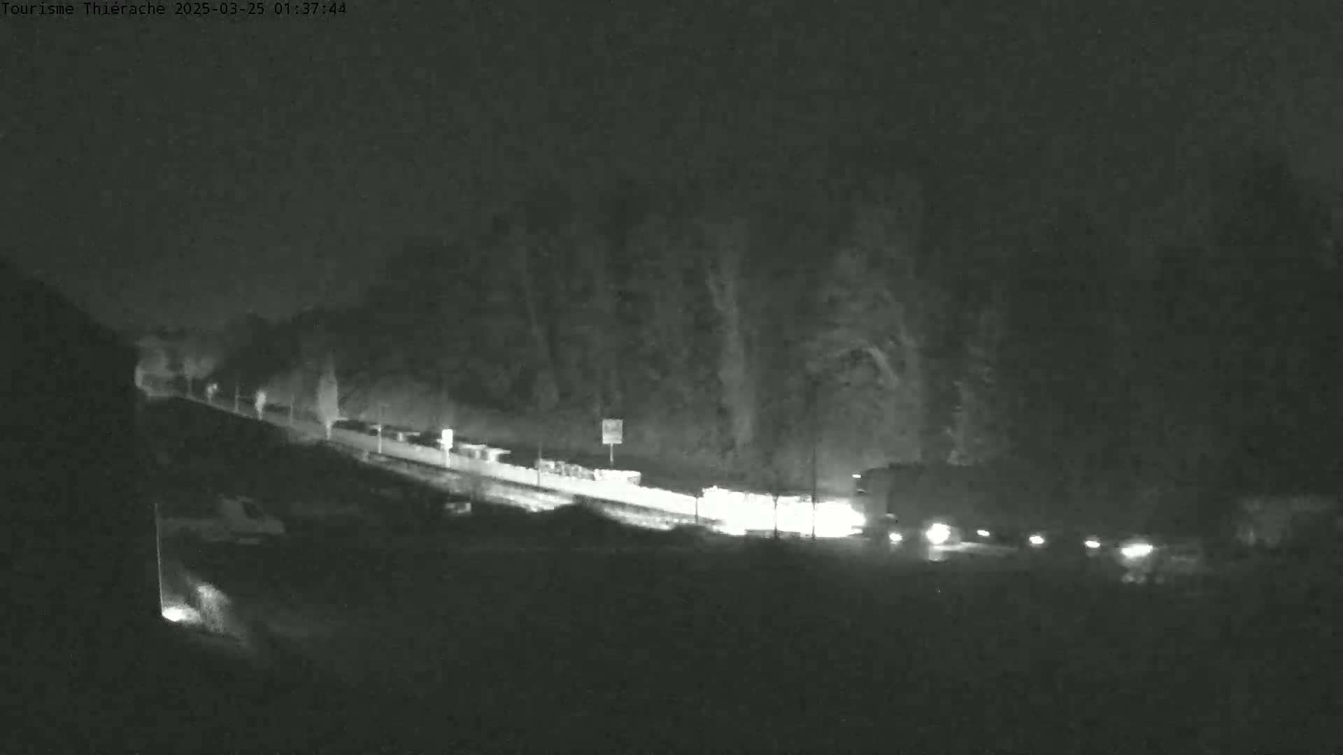 A nighttime, low-light view of a road with several vehicles traveling along a slightly sloped roadway, bordered by trees.