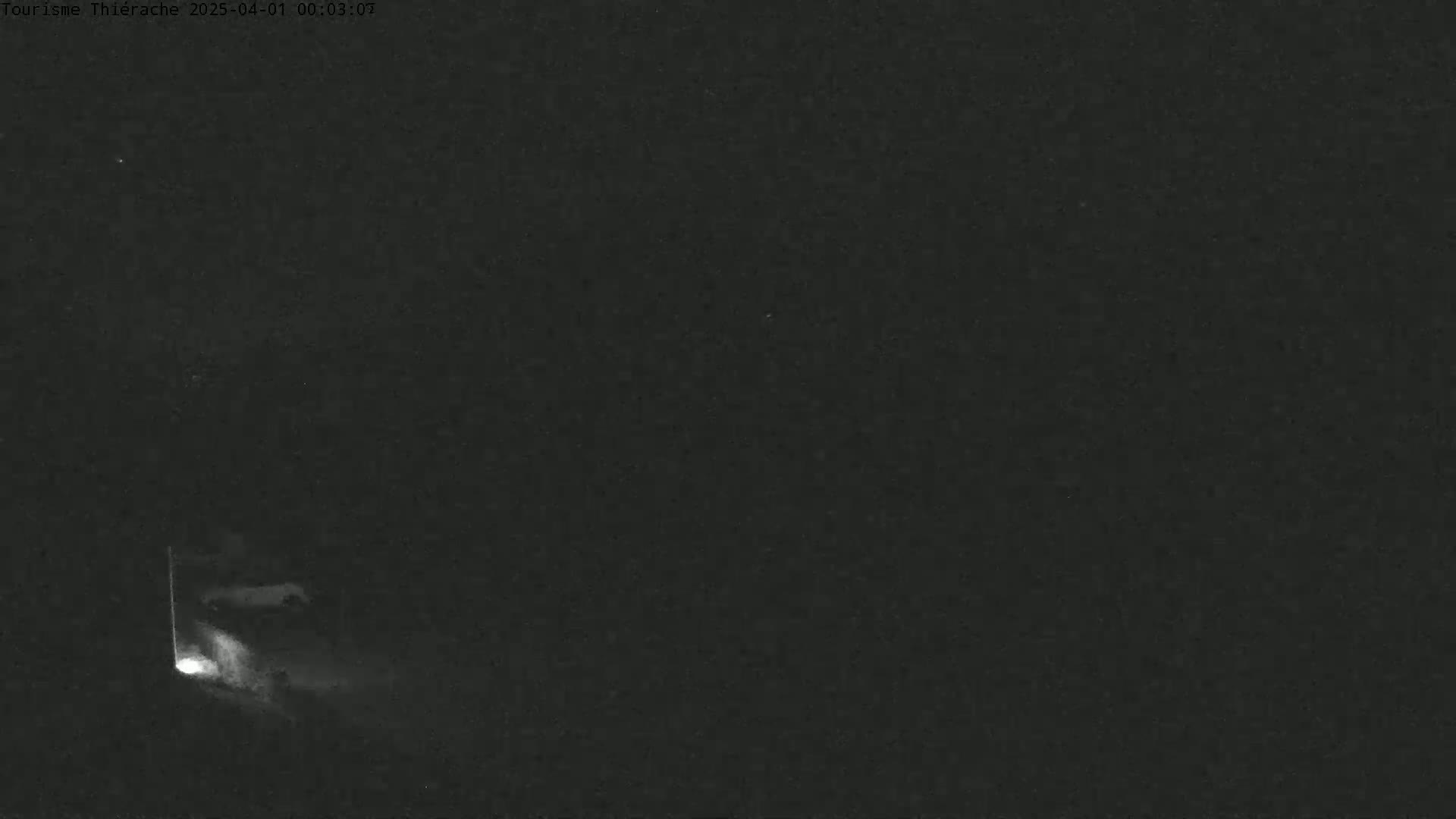 A low-light nighttime image shows a bright light source near a vertical structure, possibly a post or pole, in an otherwise dark and empty scene.
