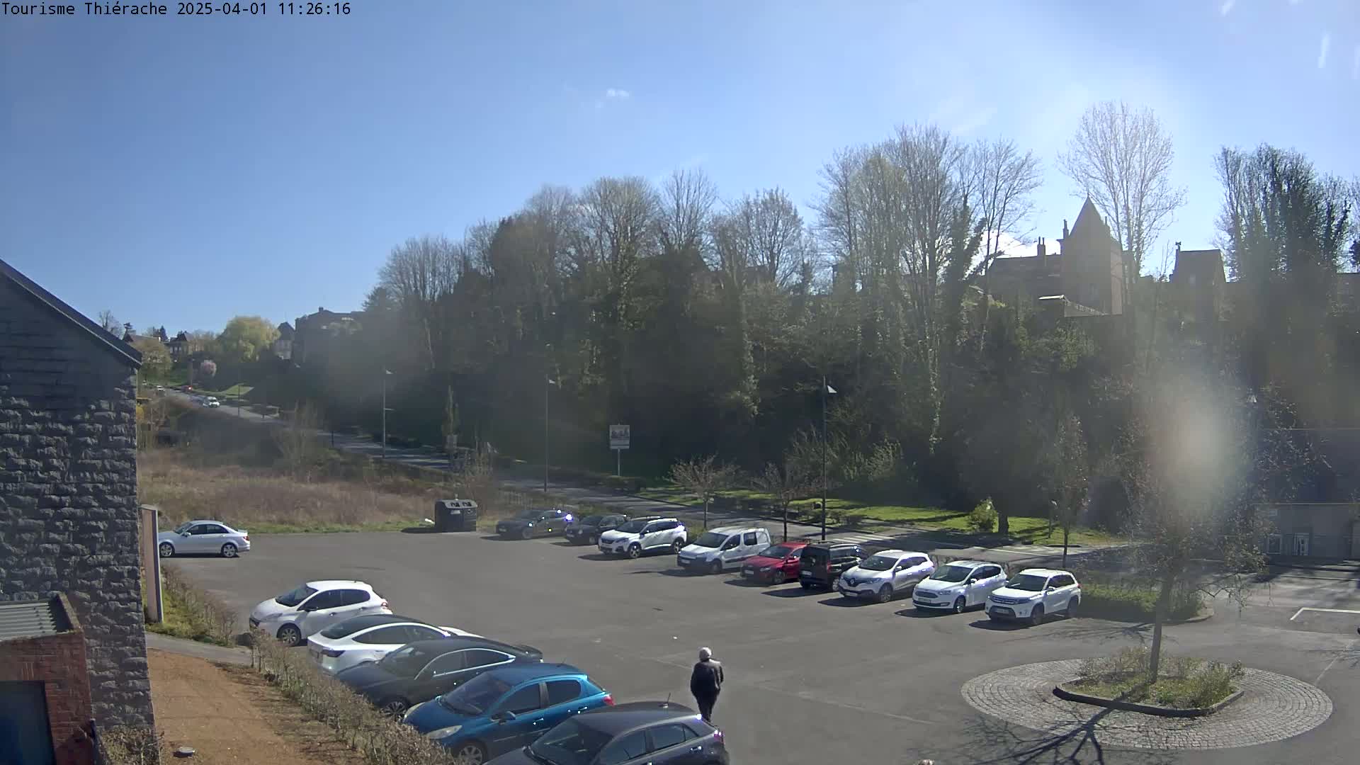 A sunny day reveals a parking lot full of cars situated below a hill covered in leafless trees and buildings.