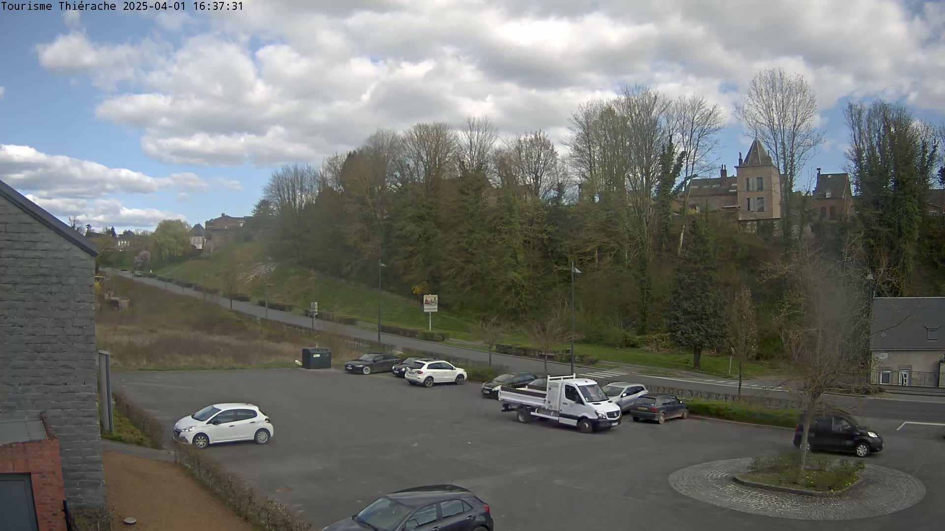 A partly cloudy day shows a parking lot with several cars and a small truck, overlooking a road leading to a hillside with buildings and trees.