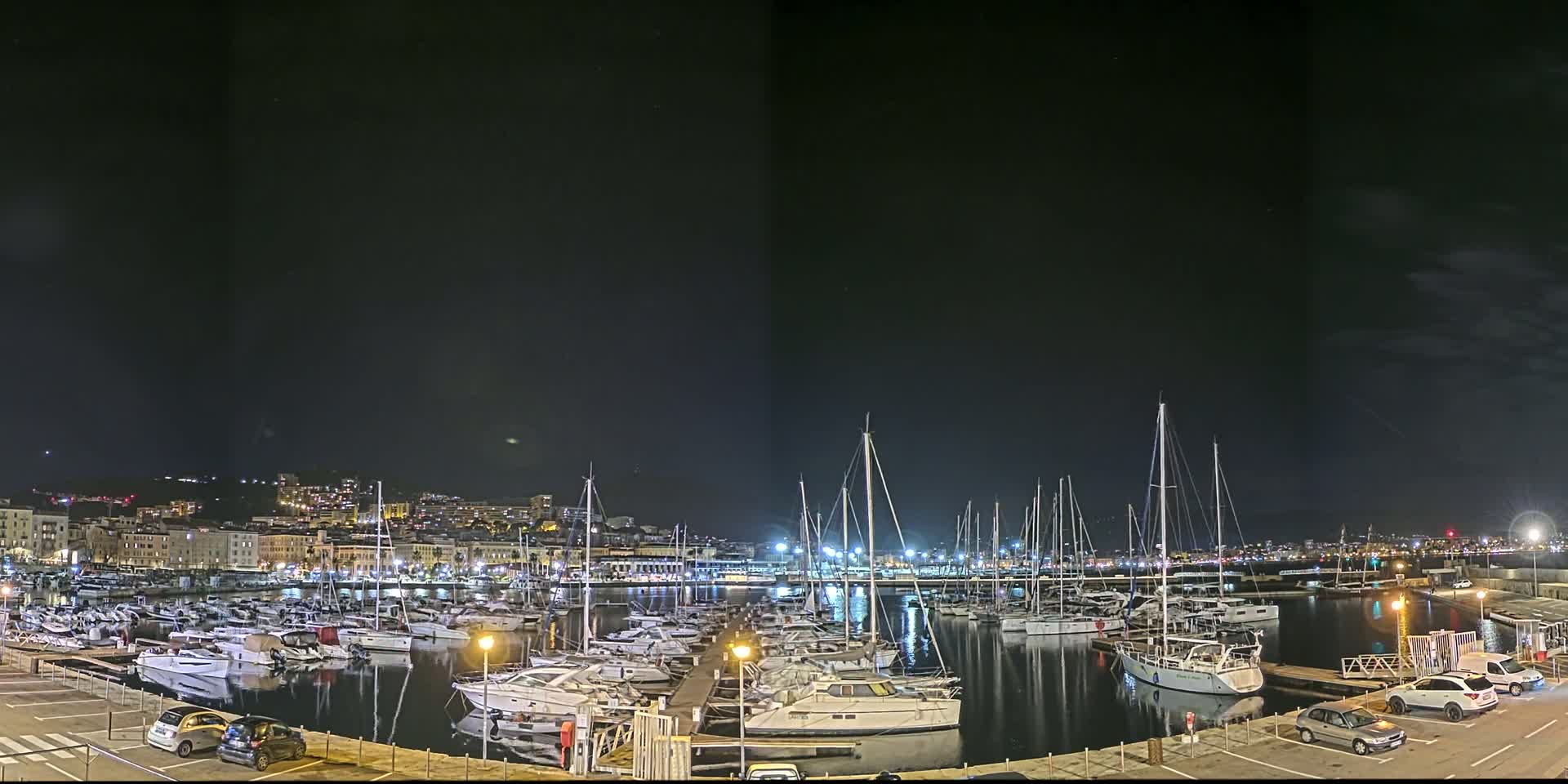 A marina filled with numerous boats is illuminated at night under a dark sky, with a city skyline visible in the background.