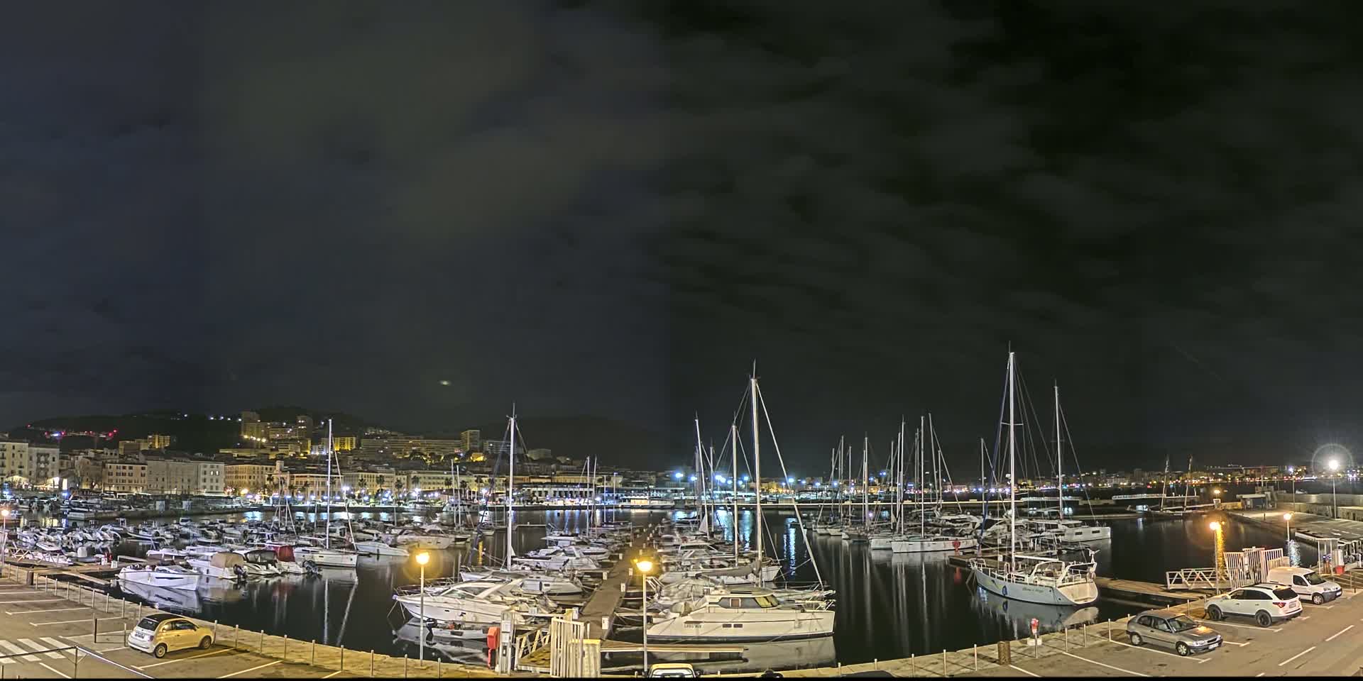 A nighttime harbor filled with many sailboats and yachts under a dark, cloudy sky.