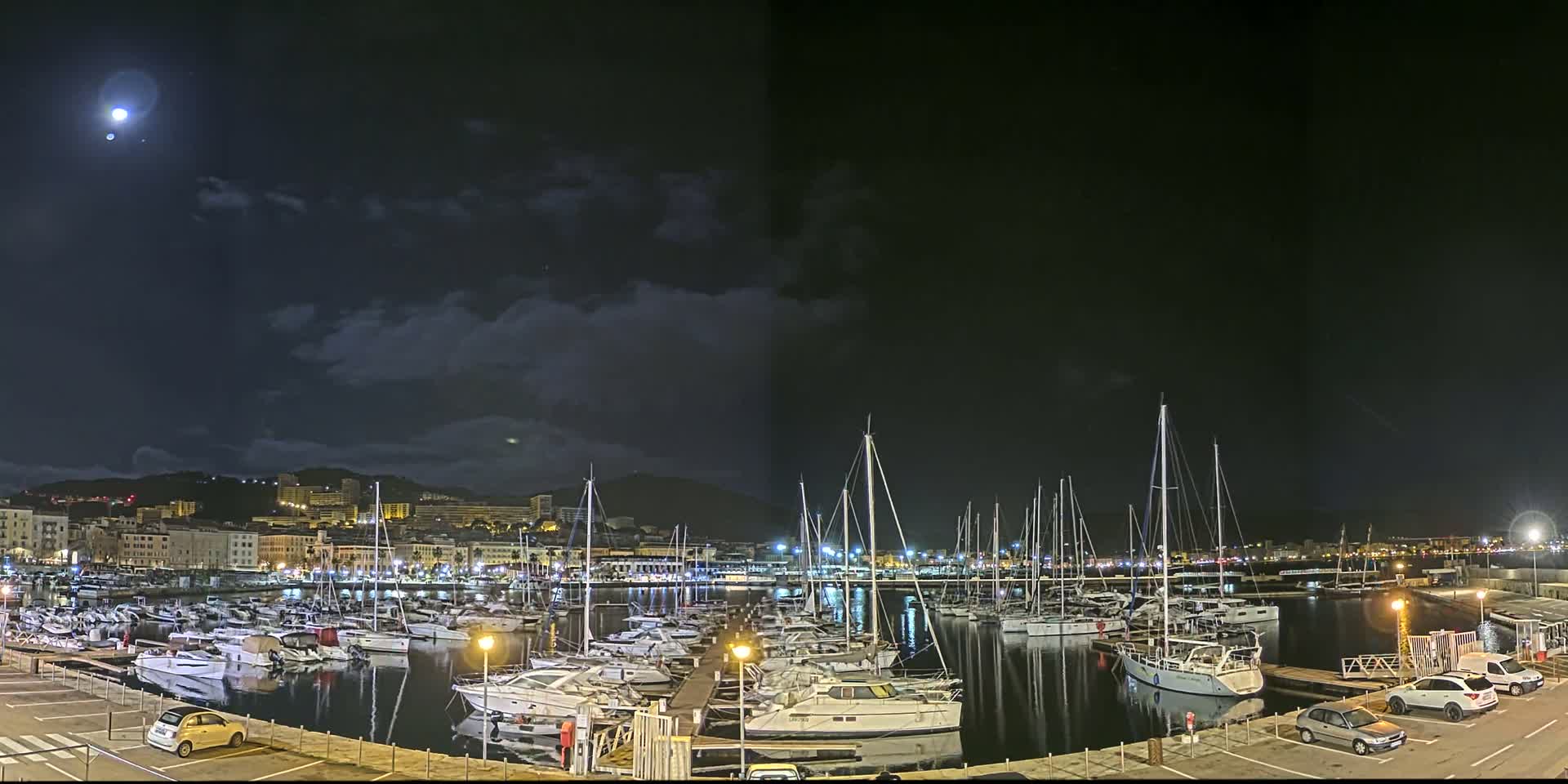 A nighttime view of a marina filled with sailboats and motorboats, under a partly cloudy sky with a bright moon.