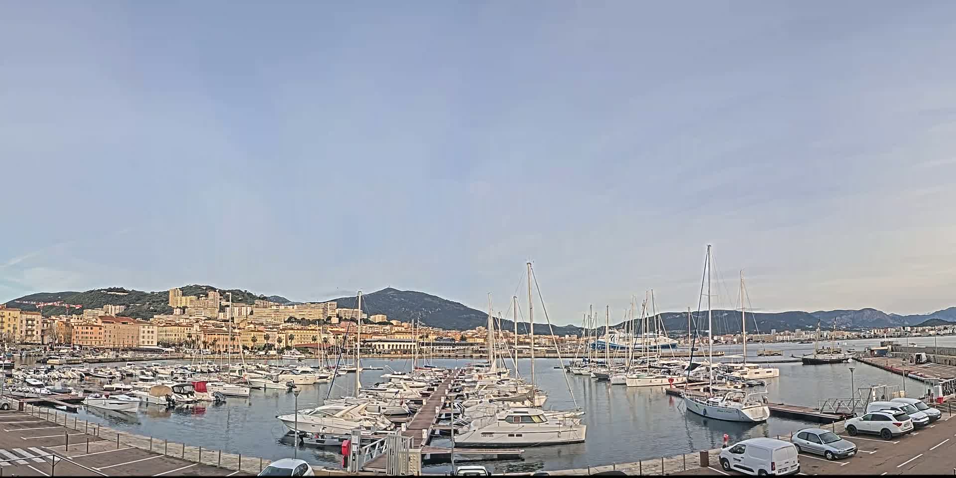 A harbor filled with sailboats and motorboats is nestled against a city backdrop under a clear, pale sky.