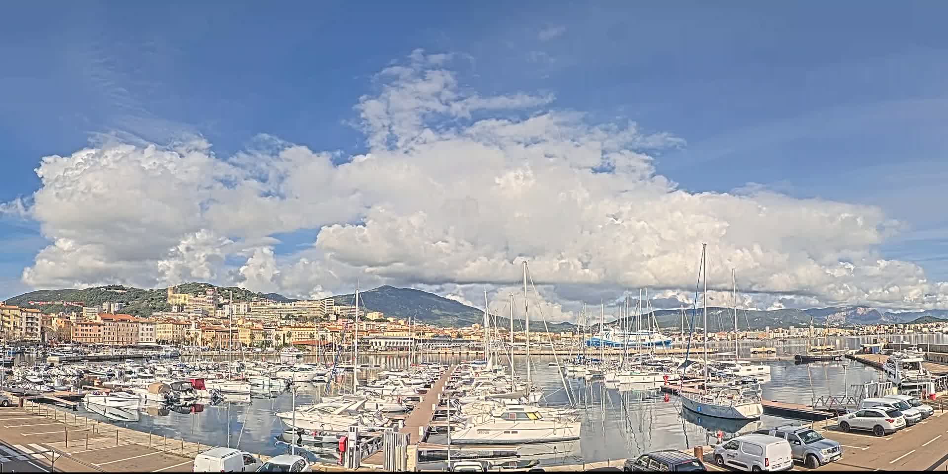 A marina filled with numerous sailboats and motorboats is situated near a city under a partly cloudy sky.