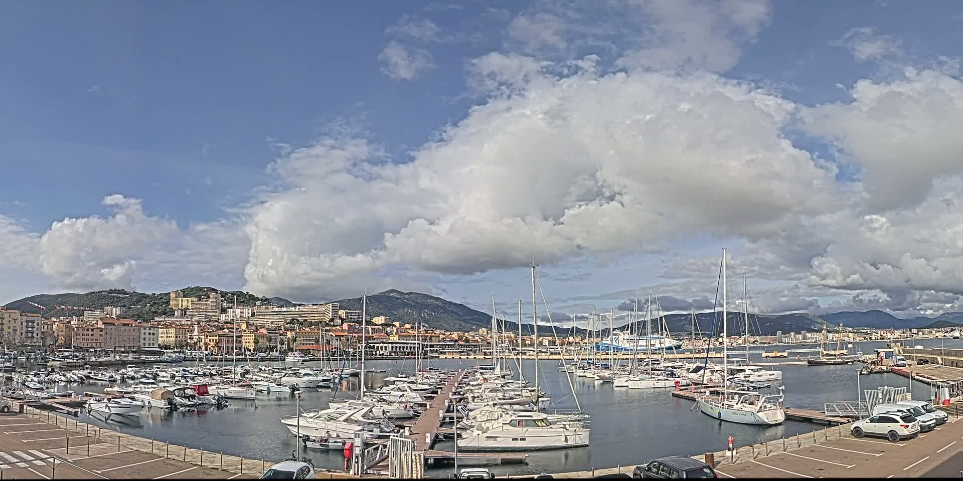 A marina filled with numerous sailboats and motorboats is situated near a city under a partly cloudy sky.