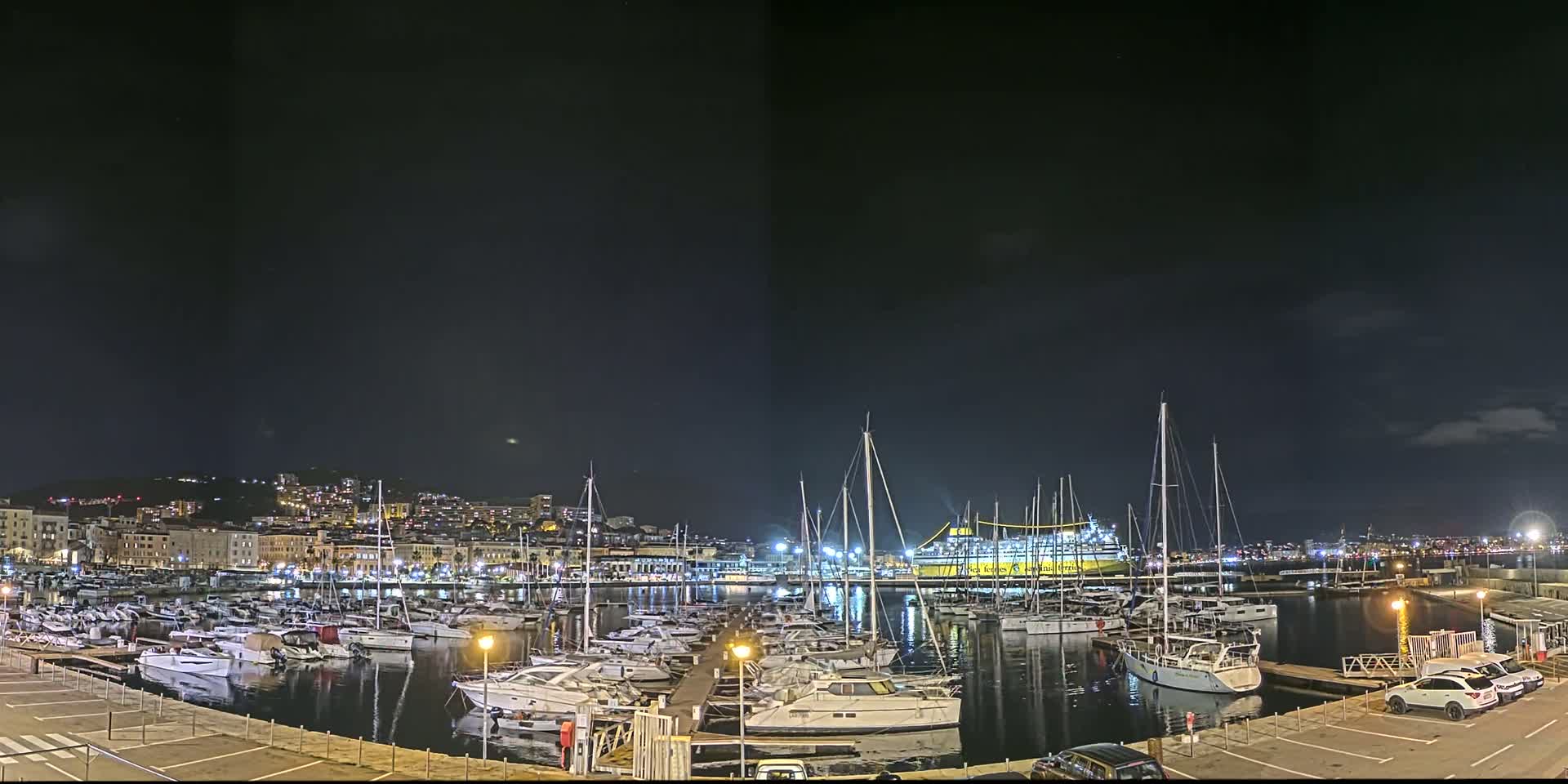 A nighttime view of a harbor filled with docked sailboats and yachts, under a dark sky with a few clouds, and a city skyline in the background.