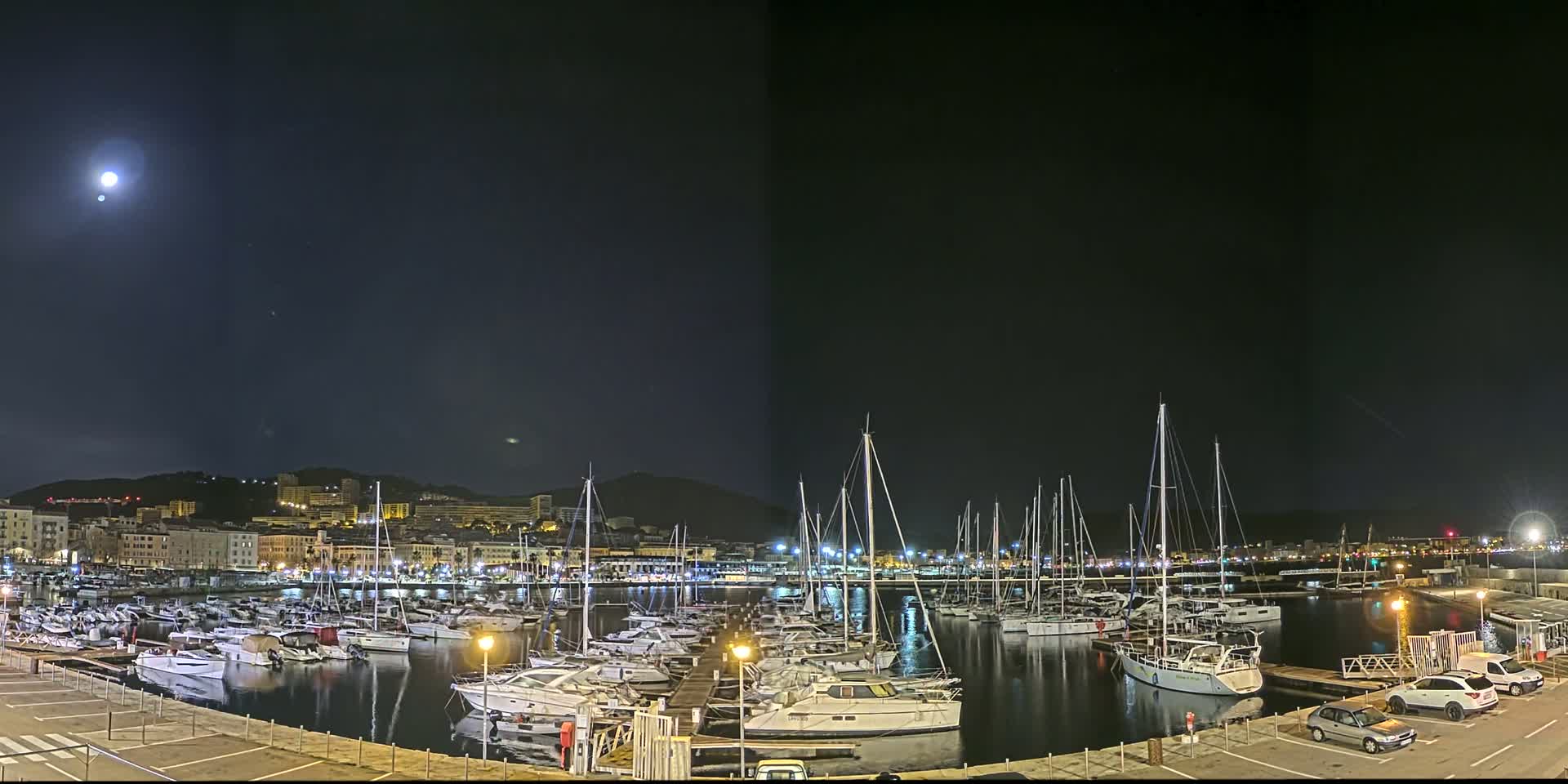 A moonlit night scene shows a marina filled with numerous boats docked, with a city and hills visible in the background.