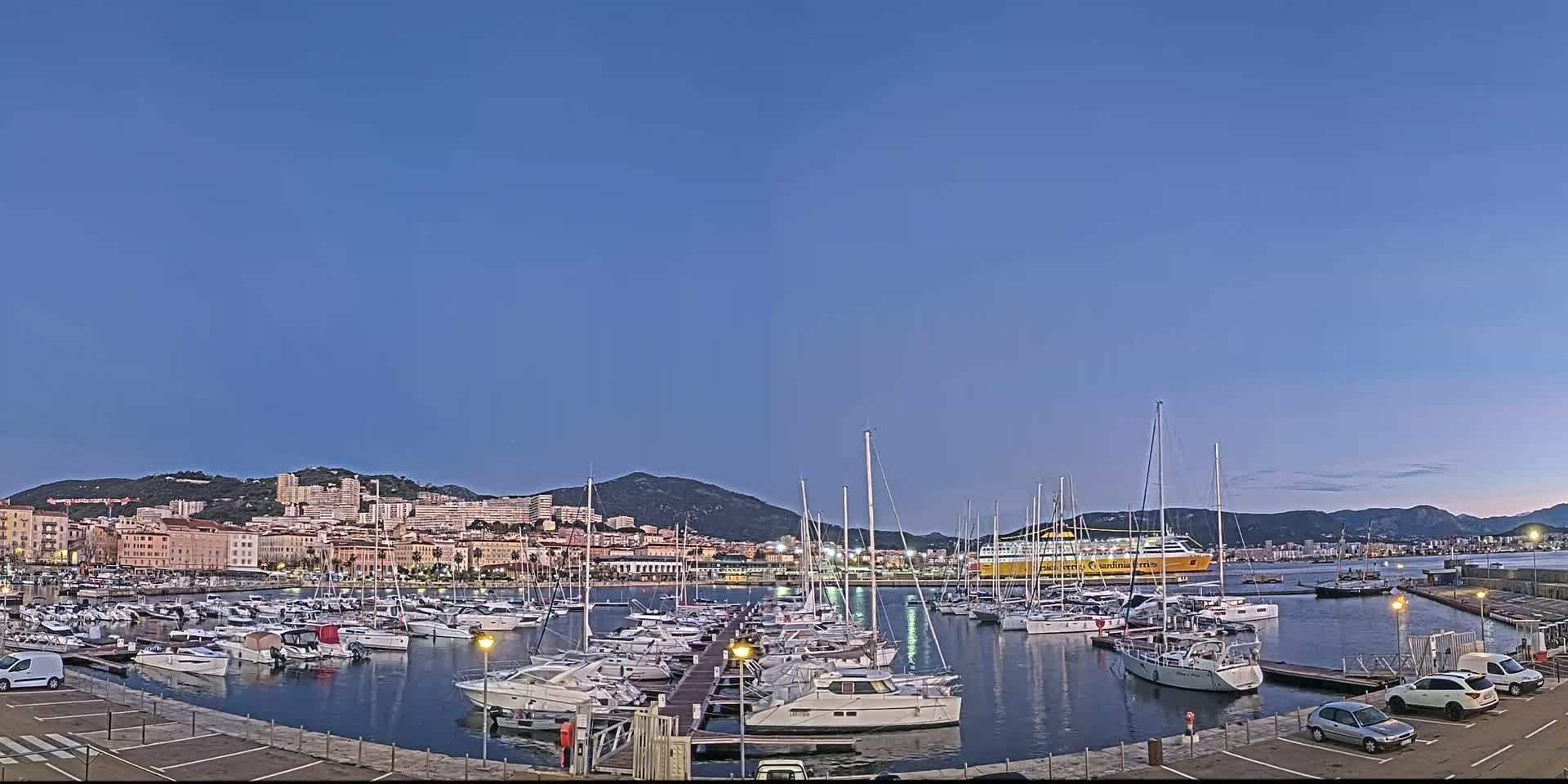 Under a twilight sky, a marina filled with sailboats and motorboats is situated in front of a city nestled against a mountain range.