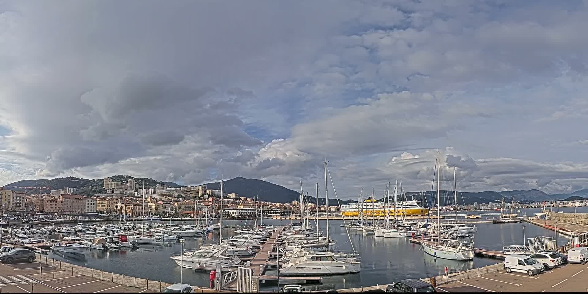 A harbor filled with numerous yachts and sailboats is situated near a city under a partly cloudy sky.