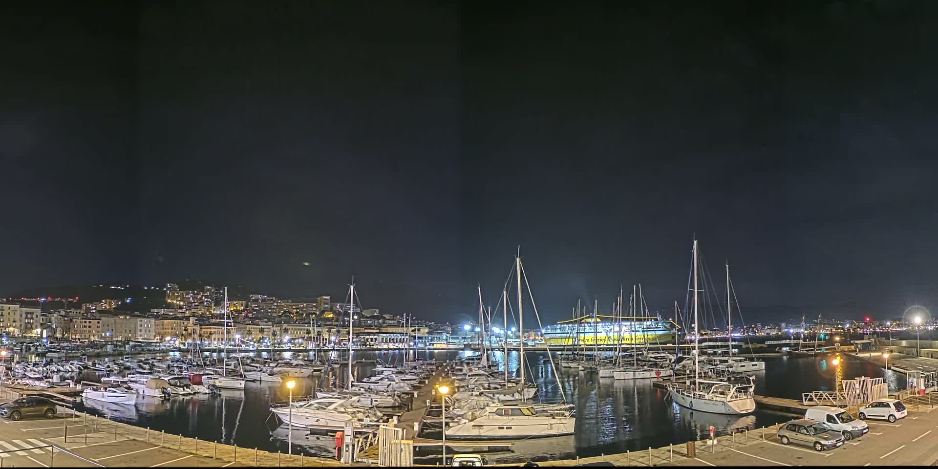 A nighttime view of a marina filled with numerous sailboats and yachts, situated in front of a city illuminated with lights under a dark, clear sky.