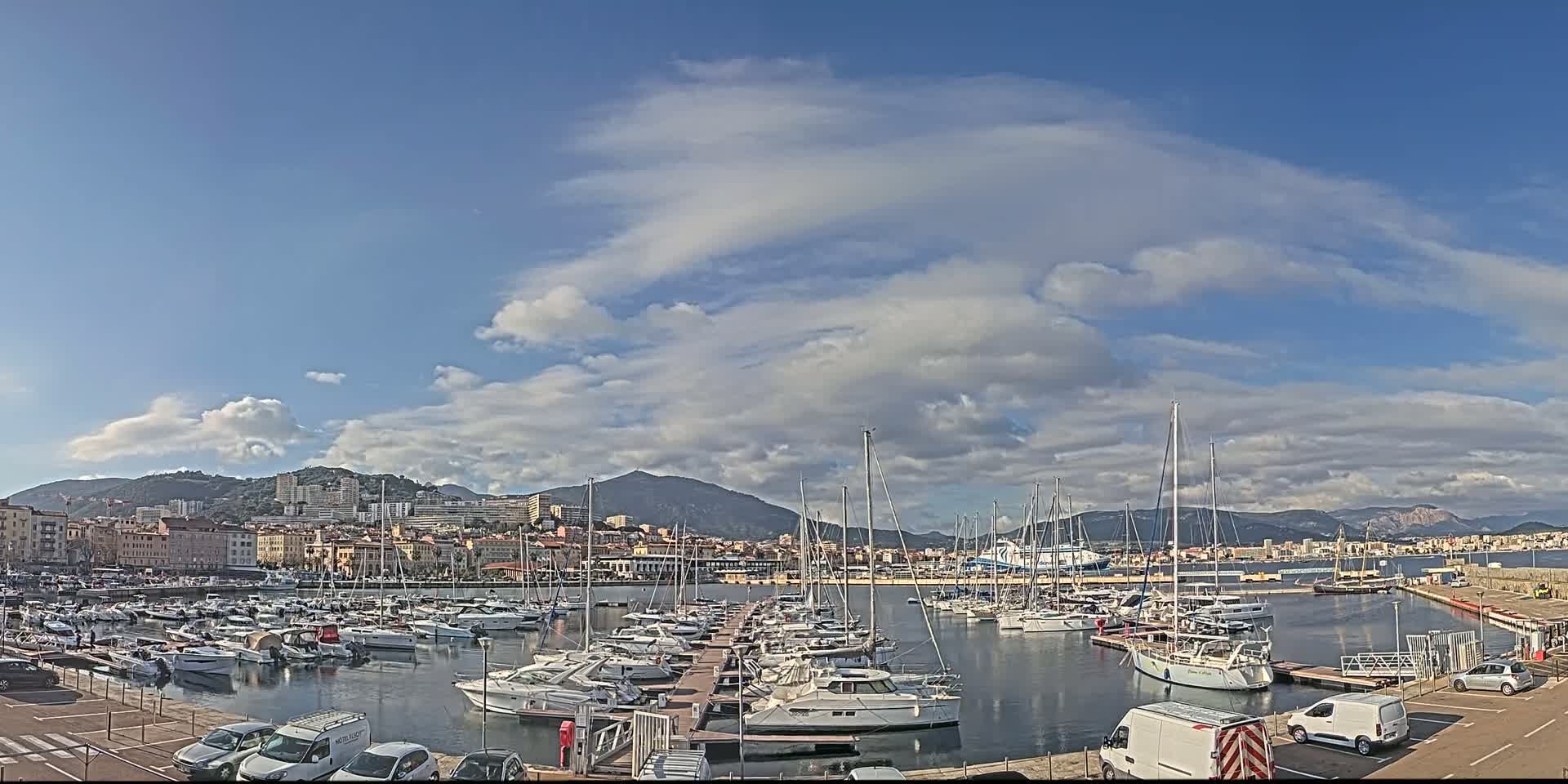 A marina filled with sailboats and motorboats is situated in front of a city and mountains under a partly cloudy sky.