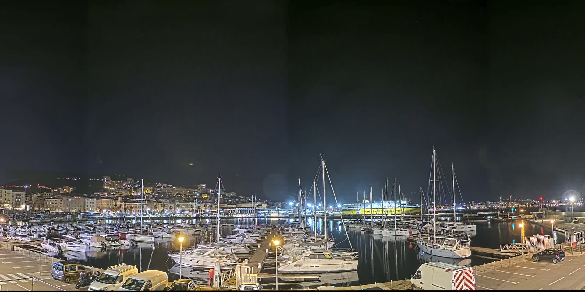 A marina filled with boats at night, under a clear dark sky, is illuminated with lights, with a city skyline in the background.