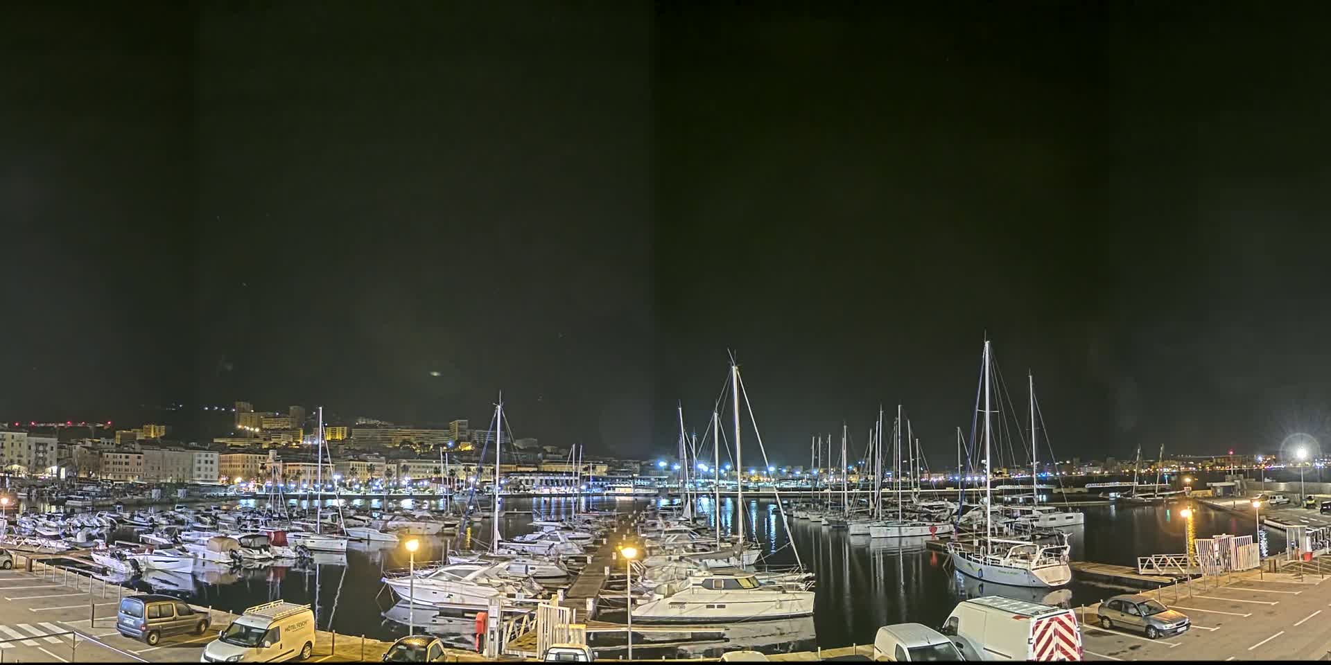 A nighttime view of a marina filled with numerous boats and yachts, under a clear, dark sky, with a city skyline visible in the background.