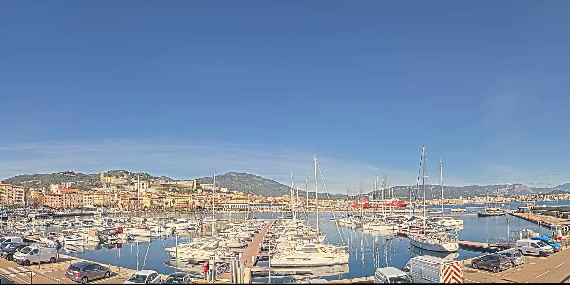A marina filled with numerous sailboats and motorboats is situated in front of a coastal town under a clear blue sky.