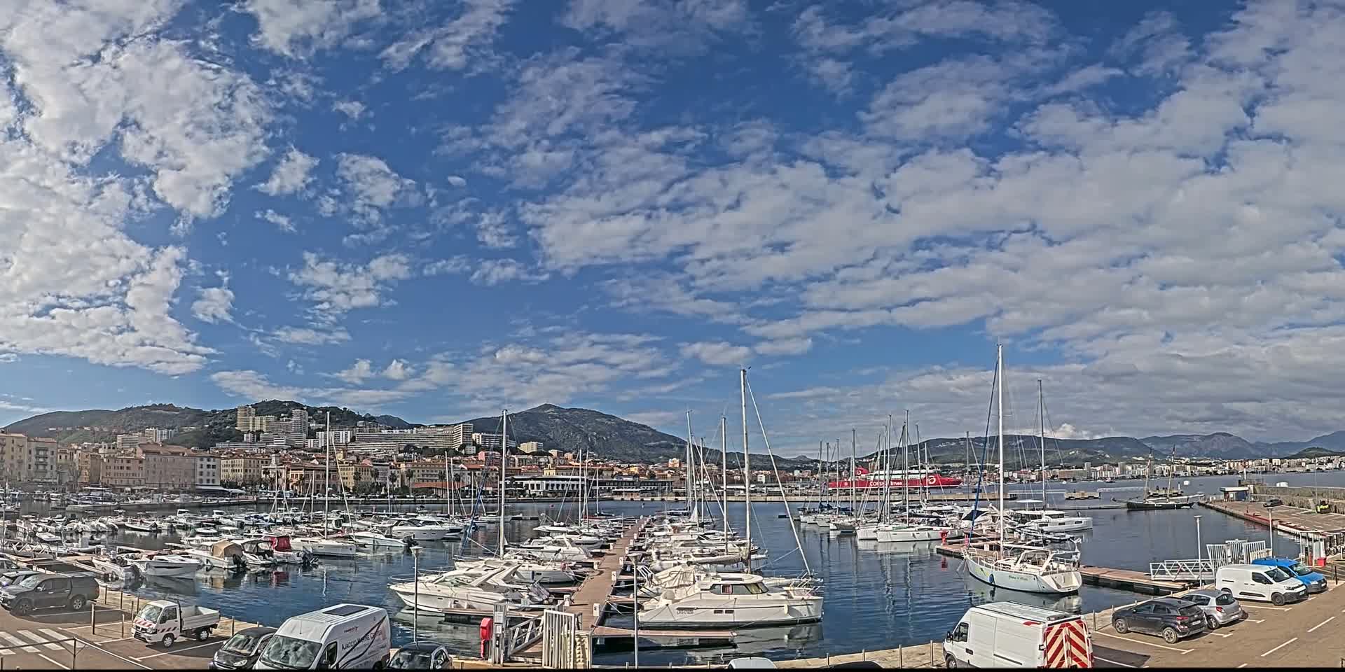 A marina filled with numerous sailboats and motorboats is situated in front of a town nestled against a mountain range under a mostly sunny sky with scattered clouds.