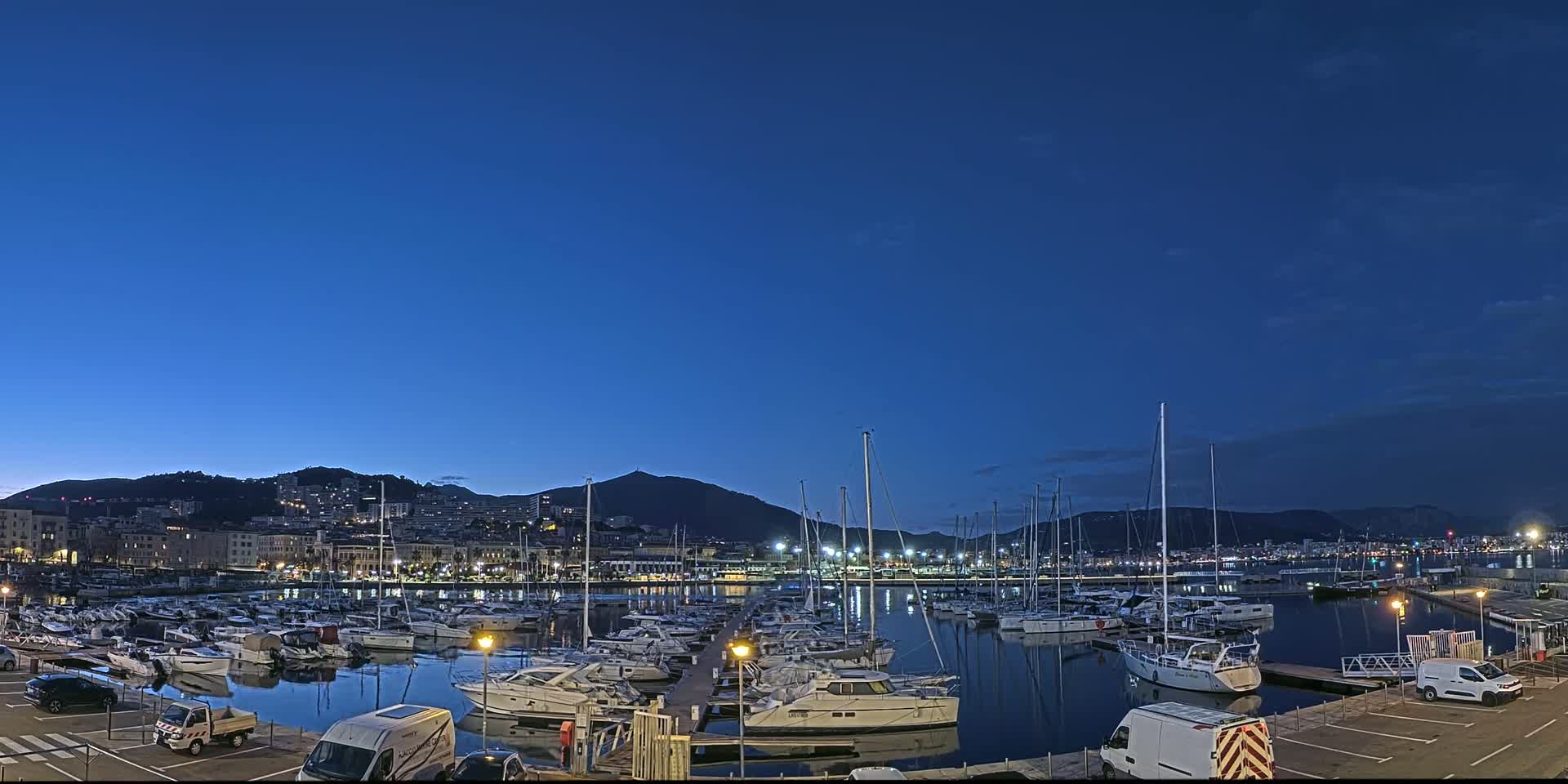 A marina filled with numerous boats is situated at twilight under a clear, dark blue sky, with a city and mountains visible in the background.