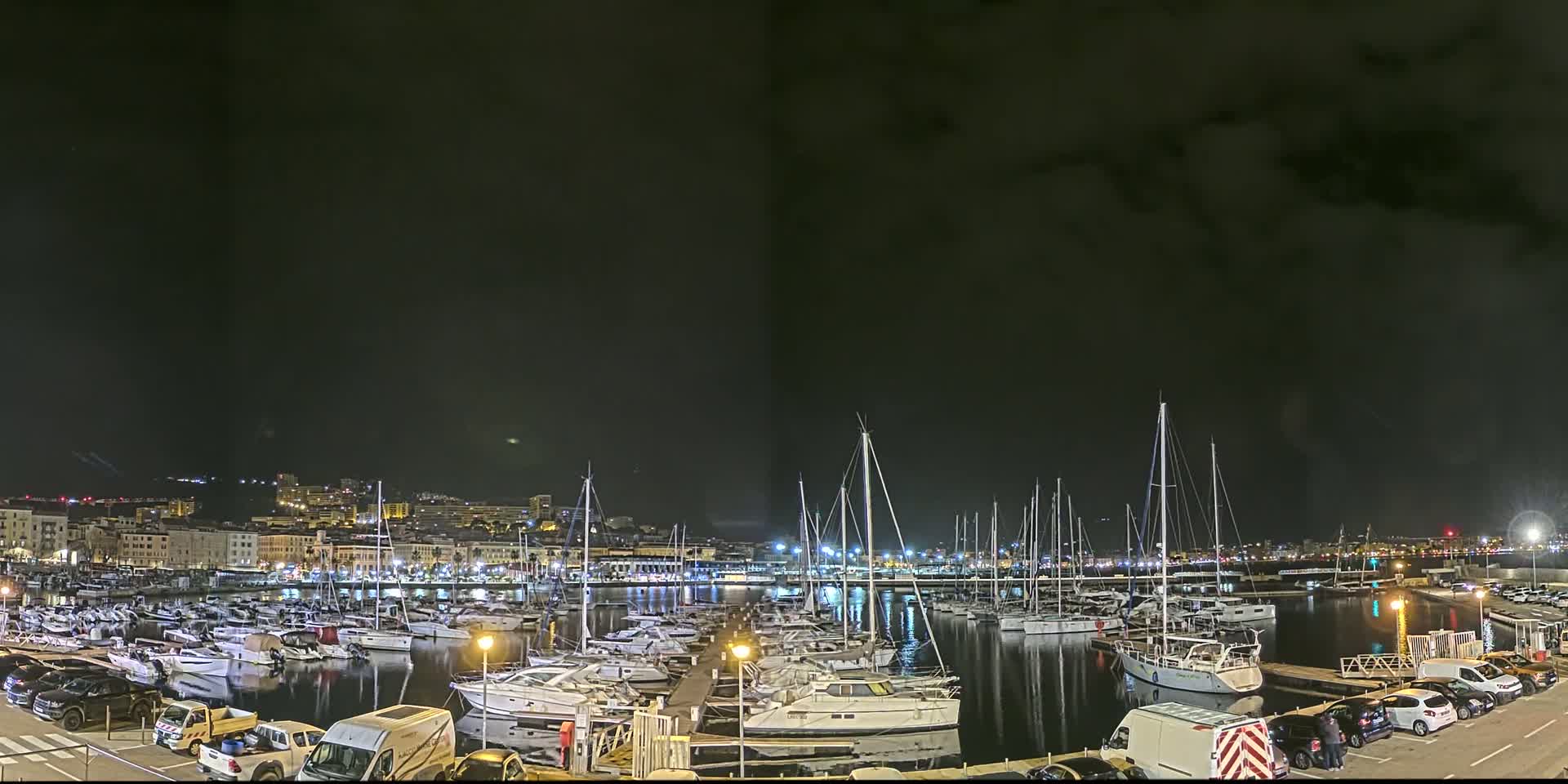 A nighttime view of a marina filled with sailboats and yachts, docked alongside a city illuminated under a dark, cloudy sky.