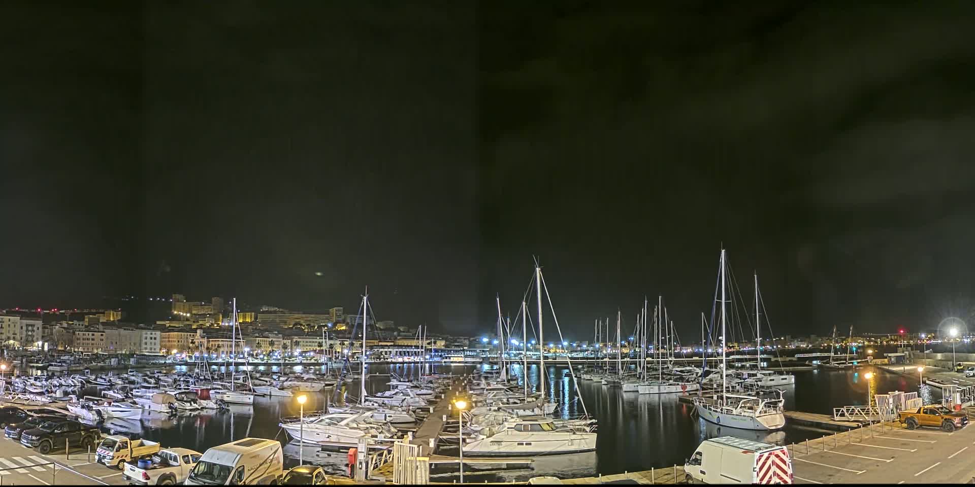 A nighttime view of a marina filled with sailboats and motorboats, under a dark, cloudy sky.