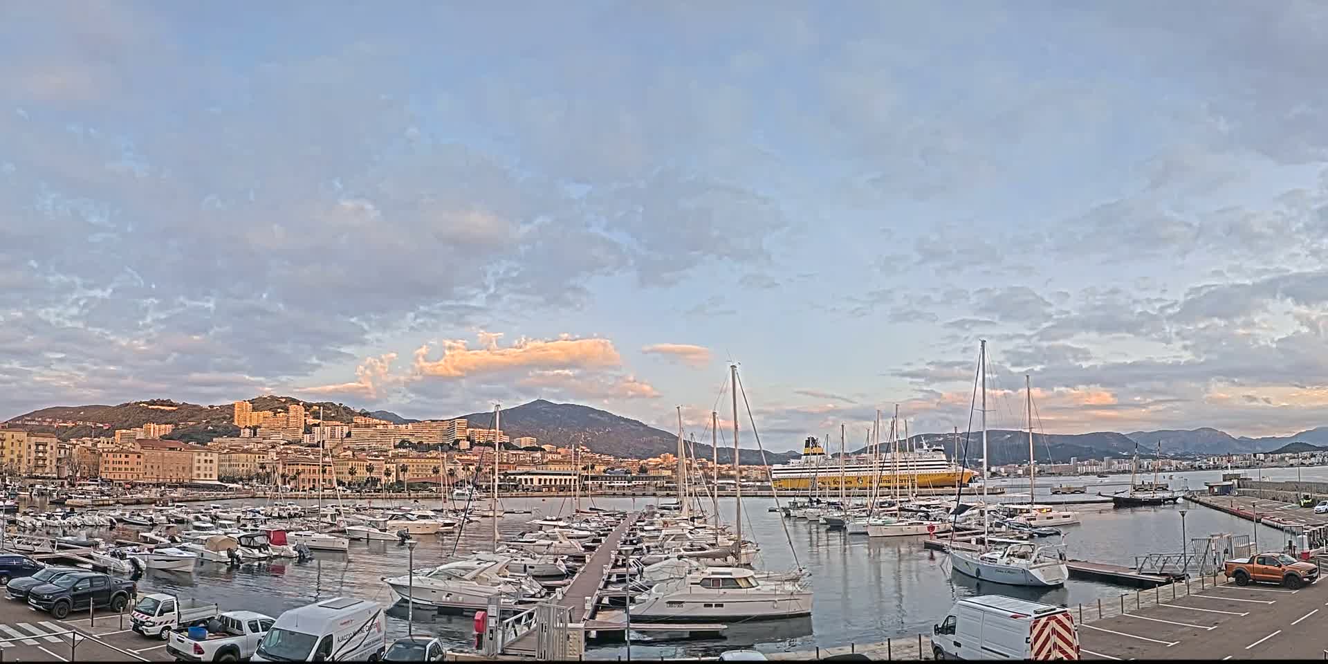 A harbor filled with numerous boats and yachts is situated in front of a city nestled against hills under a partly cloudy sky.