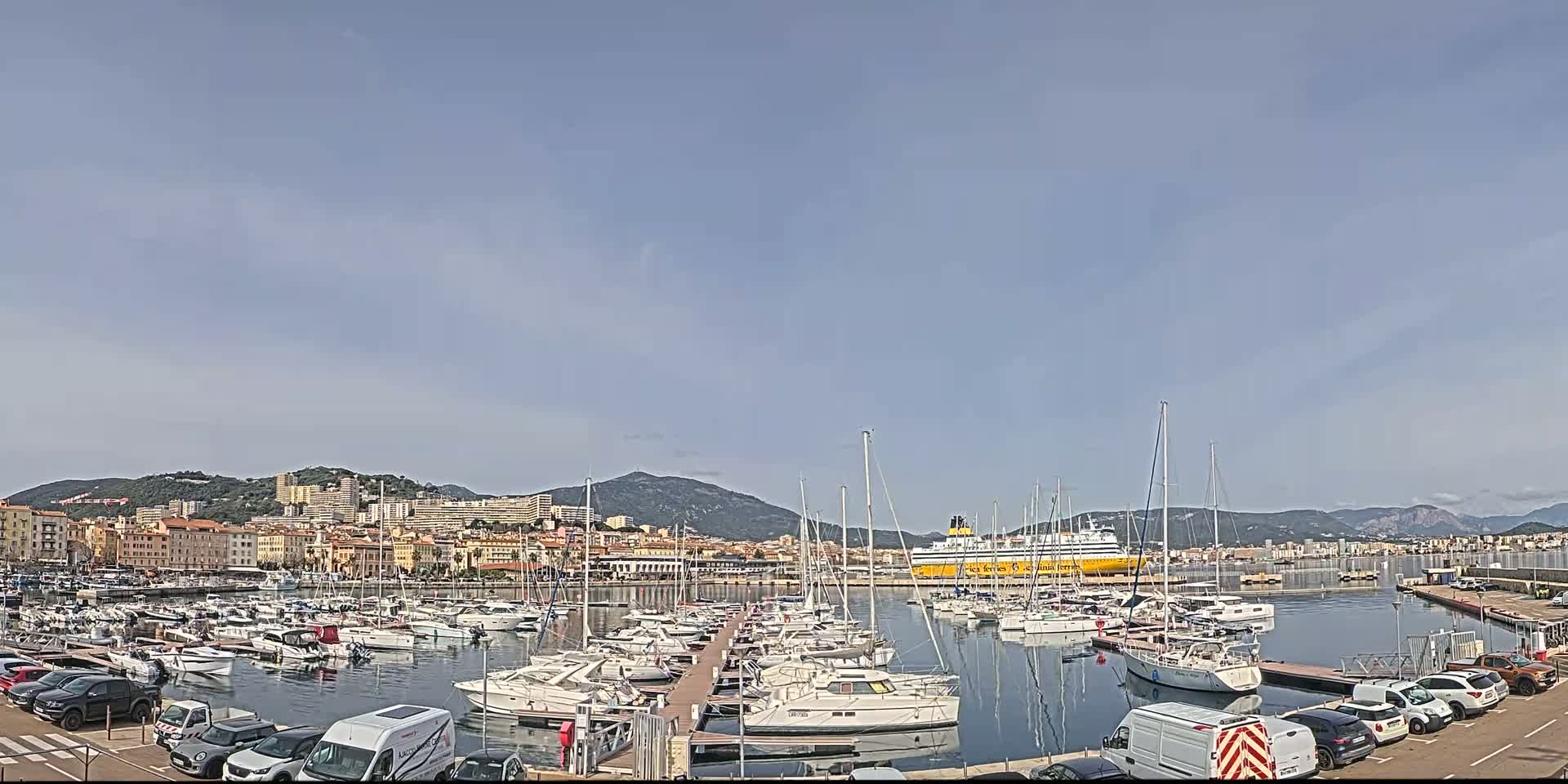 A harbor filled with numerous sailboats and motorboats is situated in front of a city nestled against a mountain range under a mostly clear sky.