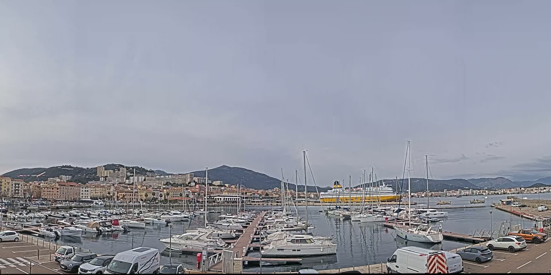 A harbor filled with numerous sailboats and motorboats is situated in front of a city nestled against a mountain range under a mostly cloudy sky.