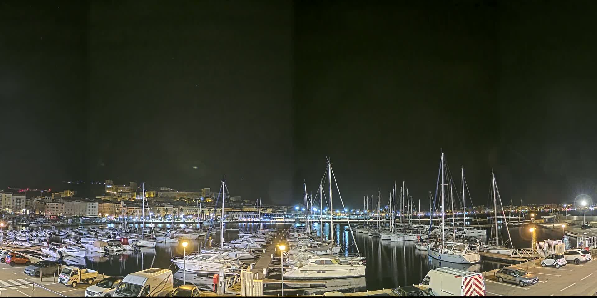 A nighttime view of a marina filled with numerous boats and yachts, docked along a waterfront city under a clear, dark sky.