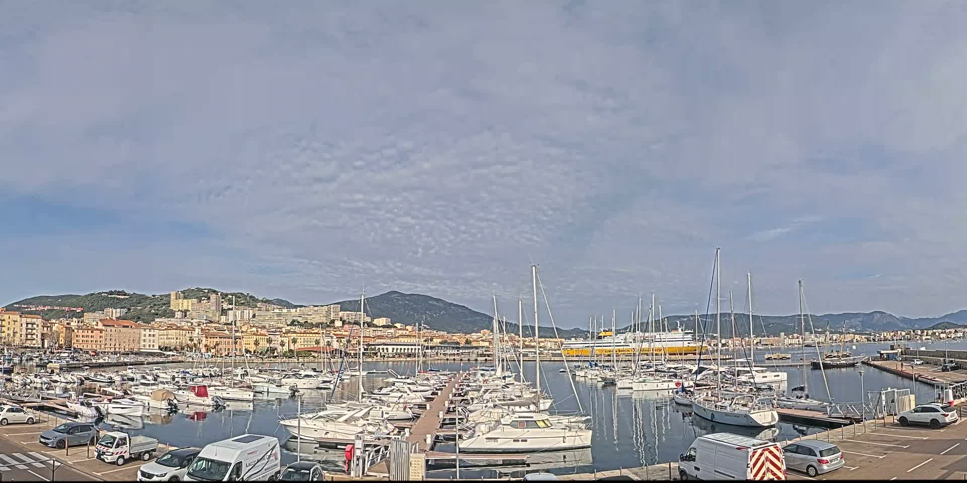 A harbor filled with numerous sailboats and yachts is situated in front of a city nestled against a mountain range under a partly cloudy sky.