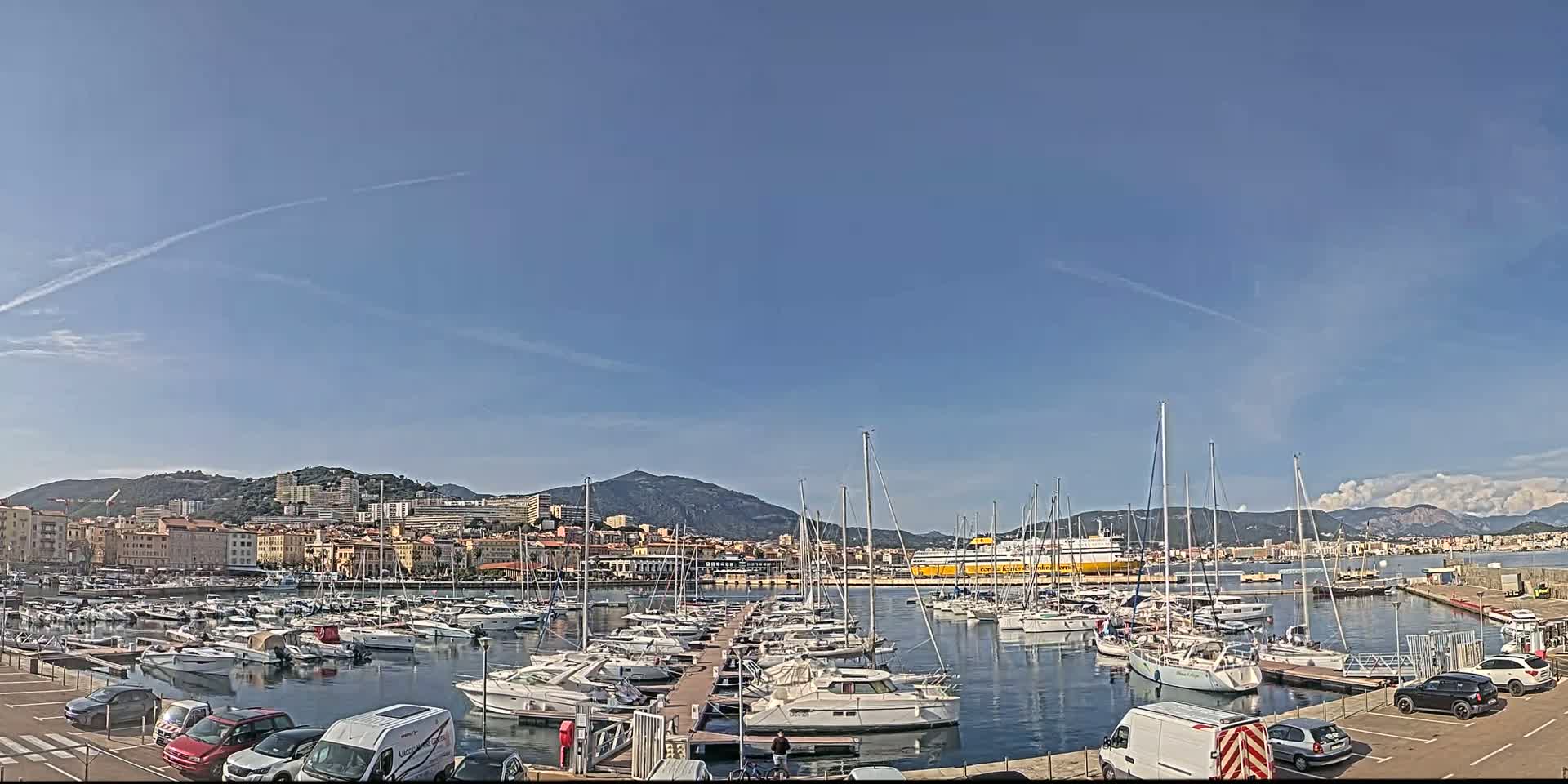 A harbor filled with sailboats and yachts is situated in front of a city and mountains under a clear blue sky.